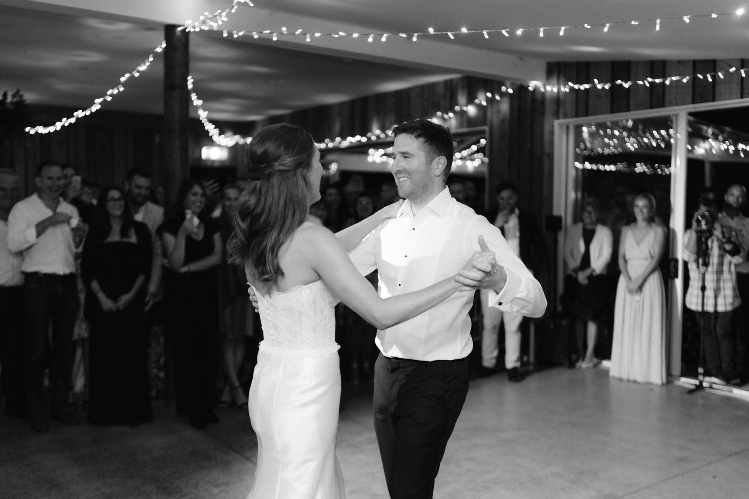 A couple dances their first dance at a wedding reception under twinkling string lights in black and white.