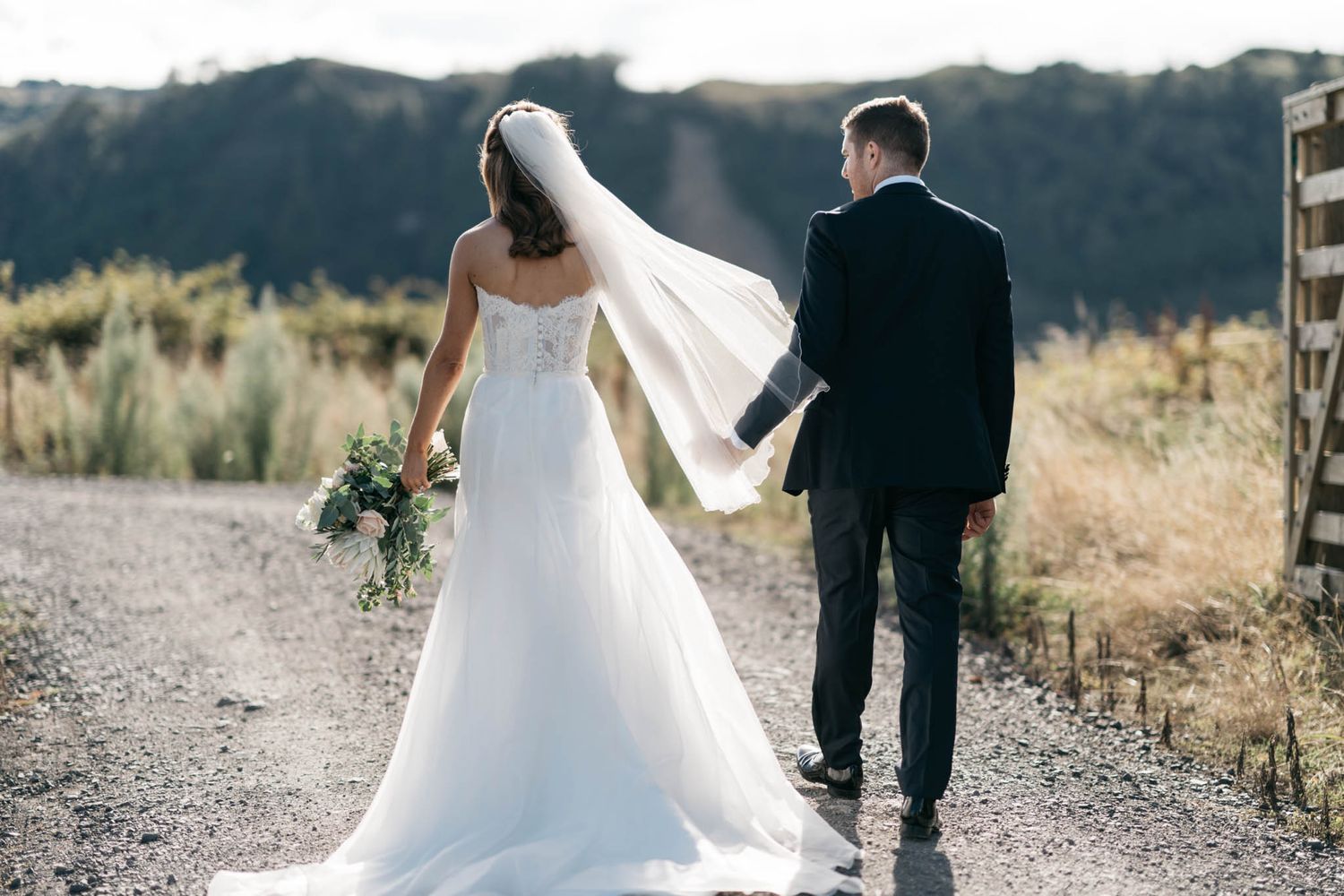 A couple walking down a dusty mountain path with the bride in a flowing white wedding dress holding a bouquet.