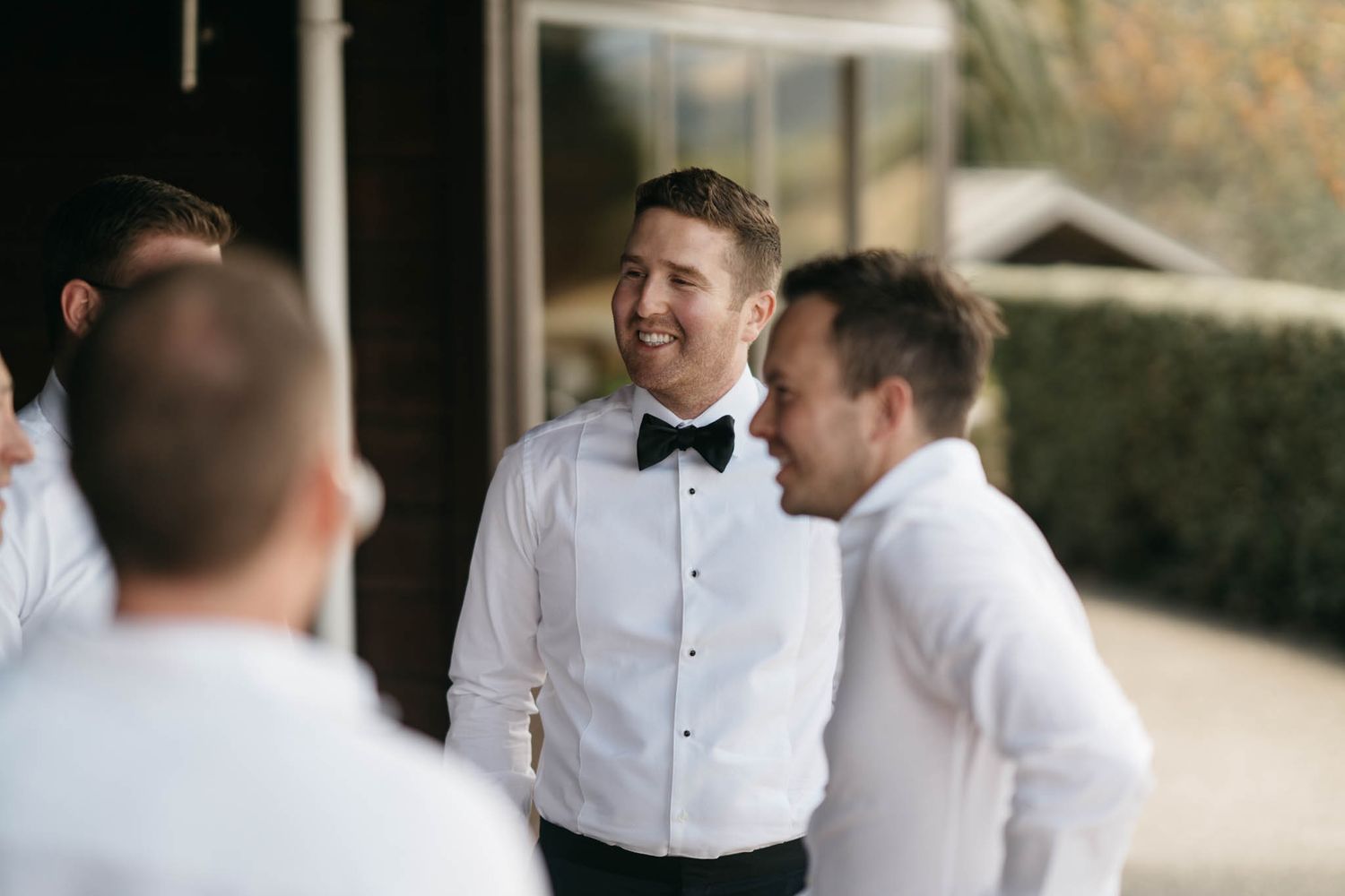 Men in white dress shirts and bowties share a candid moment while getting ready for a formal event.
