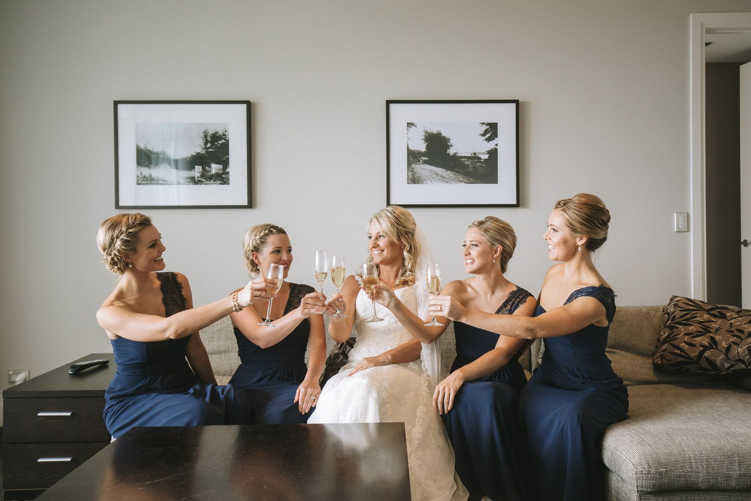 A row of bridesmaids wearing navy blue dresses sit together on a couch laughing and smiling together.