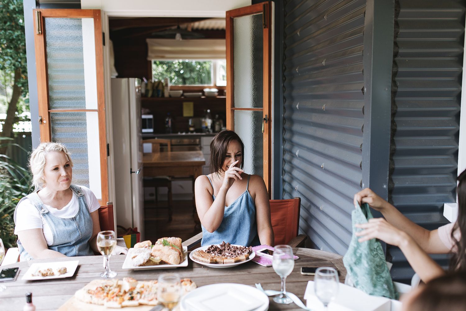 Three friends gather around a dining table on a covered porch enjoying a casual meal together on a sunny afternoon.