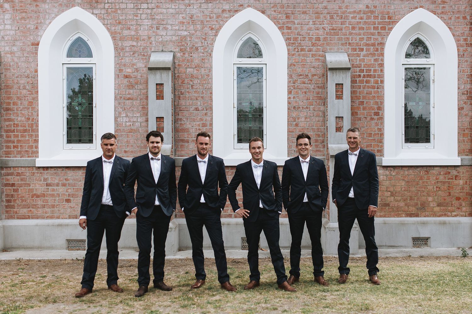 Groomsmen in black suits and ties stand confidently in front of a historic brick church with white arched windows.