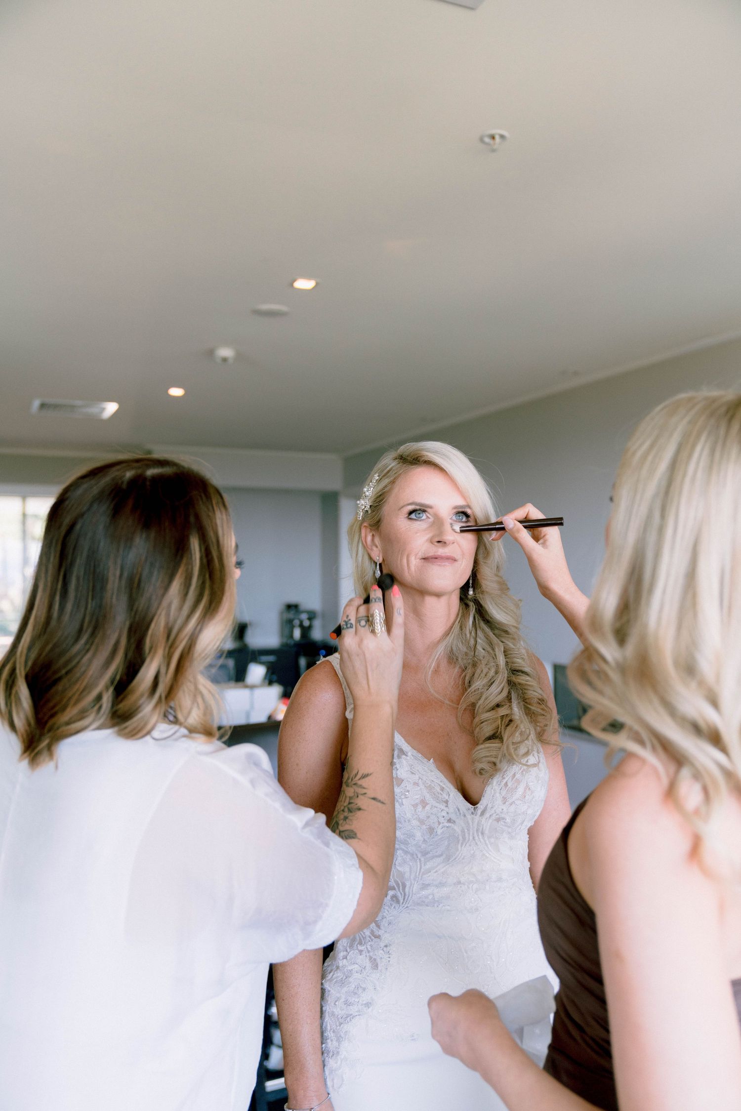 Behind the scenes bridal preparation shows stylists doing hair and makeup in a bright room.