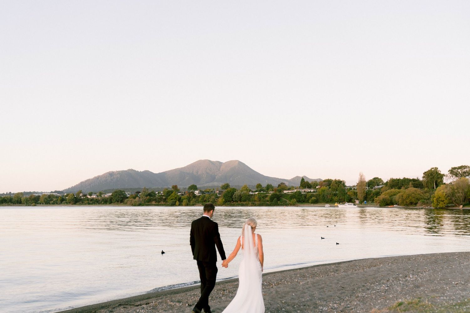 Couple walks along a peaceful lake shore at sunset with mountains in the distance.