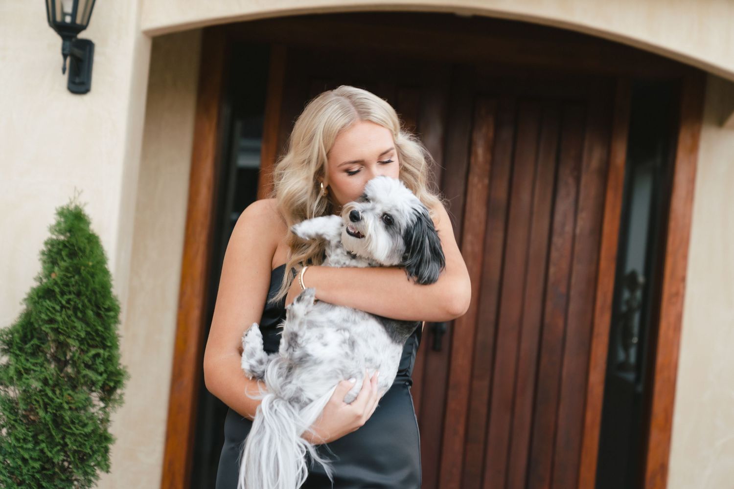 A person in black hugs a fluffy white senior dog outside a wooden door with greenery in the background.