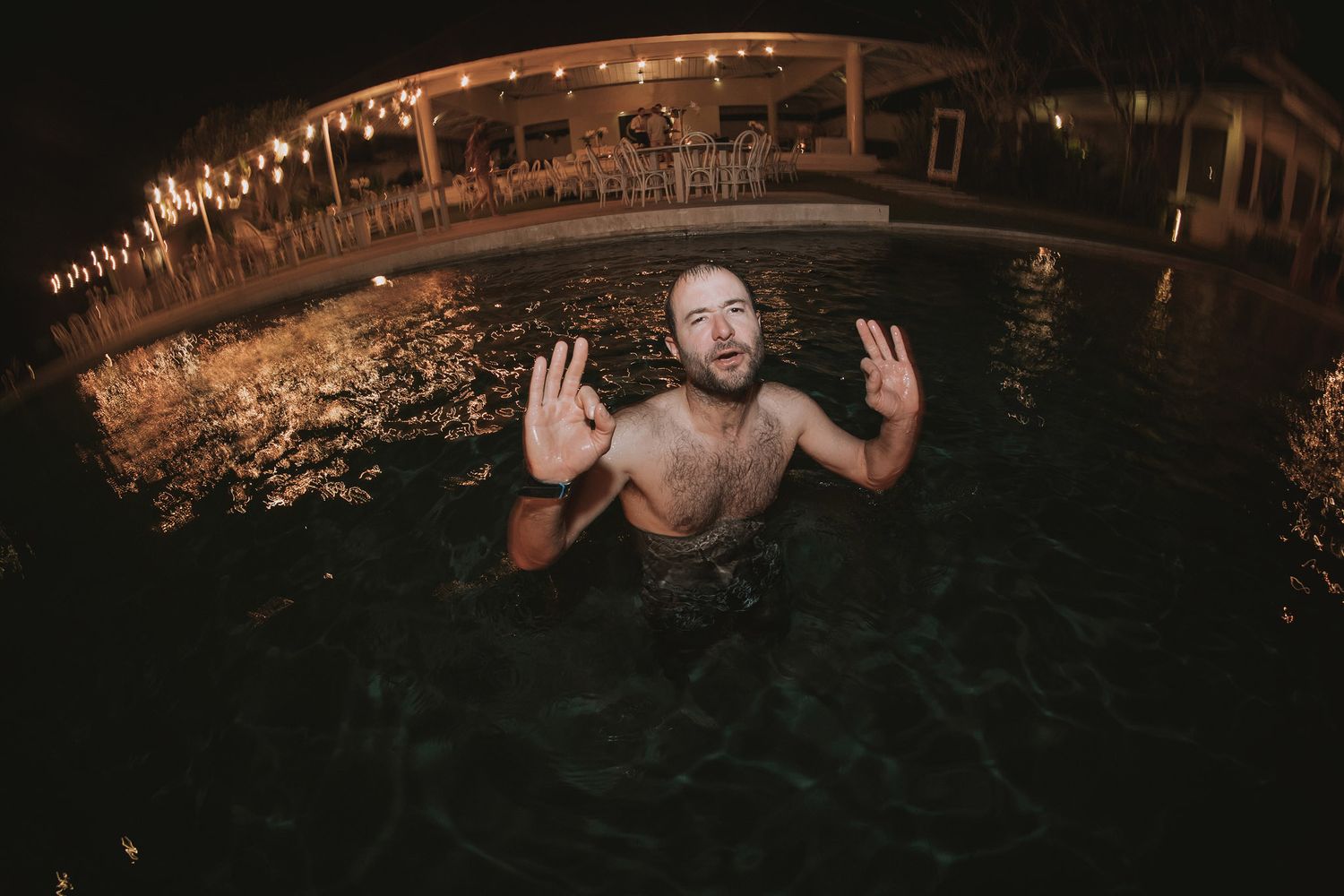 Someone playfully poses with raised hands in an illuminated swimming pool at night with twinkling lights in the background.