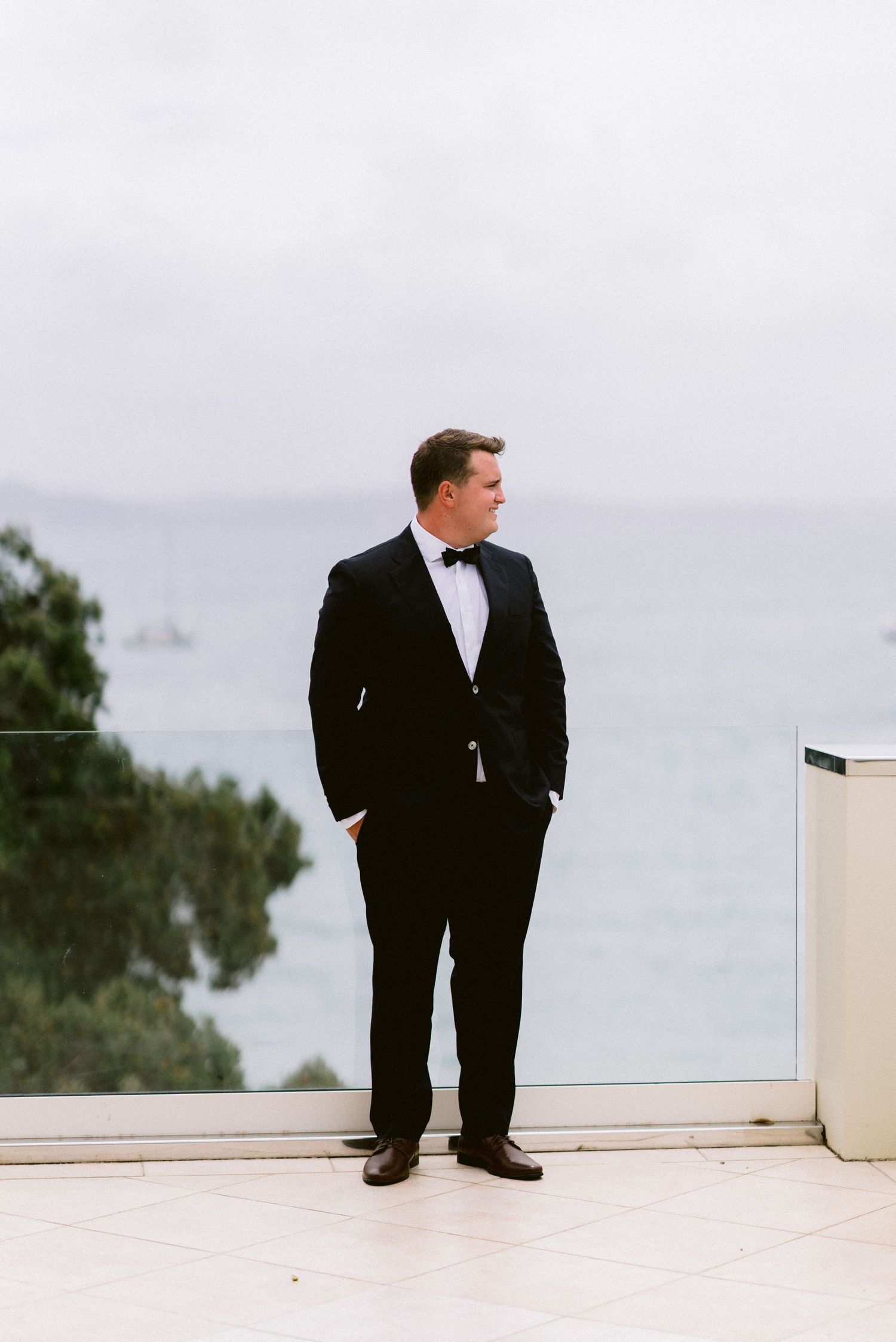 A groom in a classic black tuxedo stands contemplatively on a white balcony overlooking the ocean.