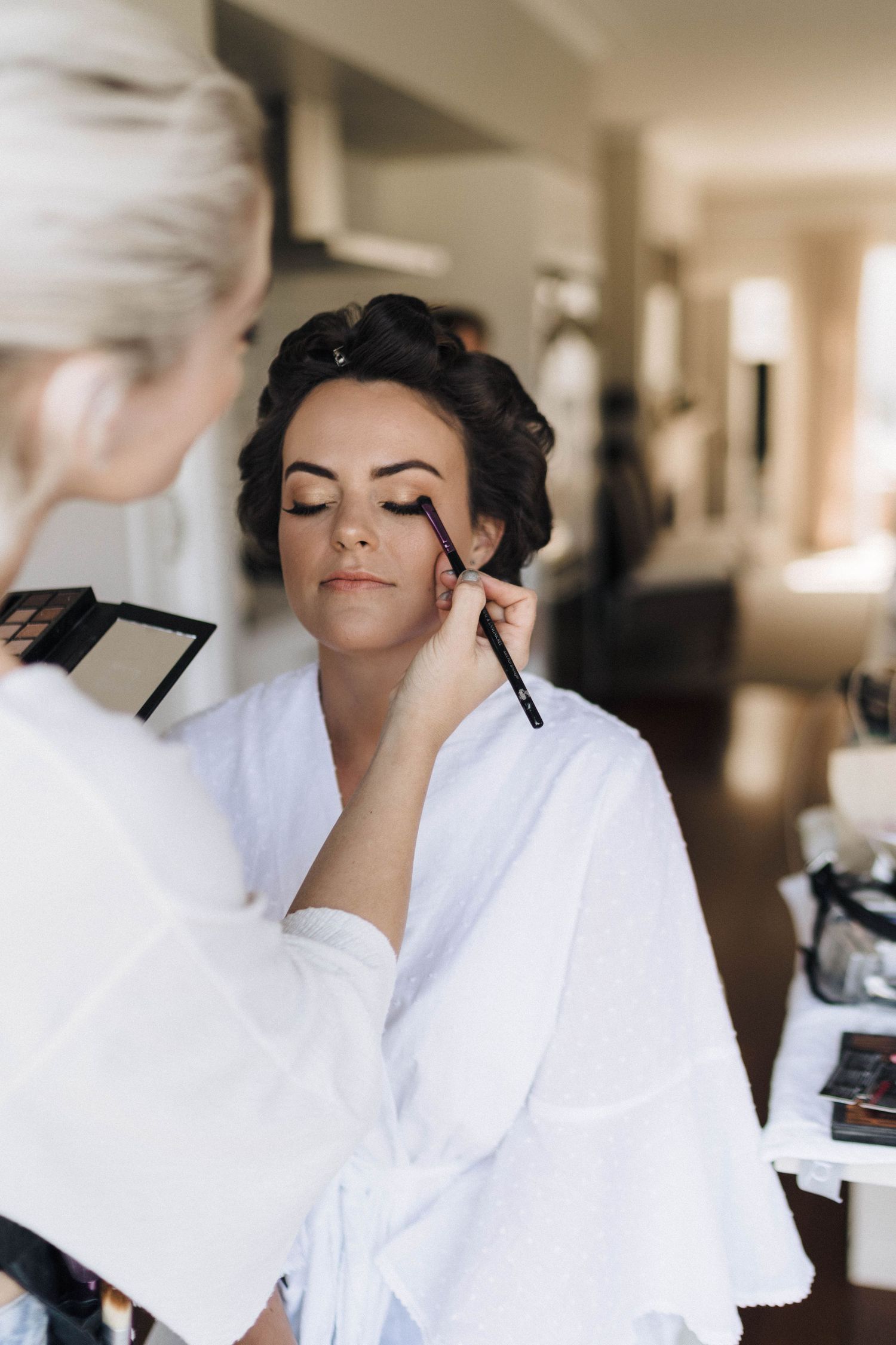Professional makeup artist applying eye makeup to client wearing white robe in bright studio setting.