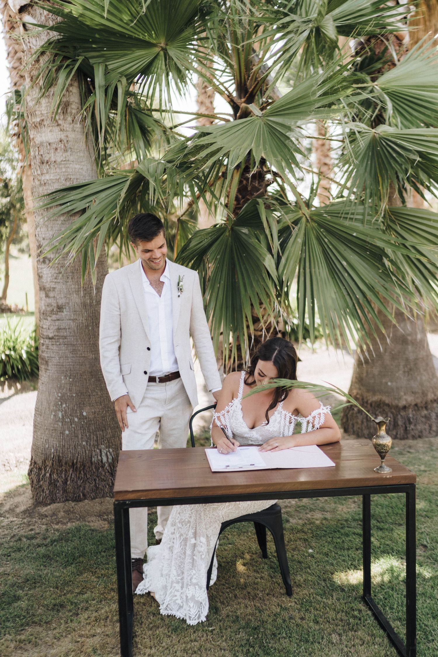 Wedding couple signing documents at rustic wooden table under tropical palm trees.