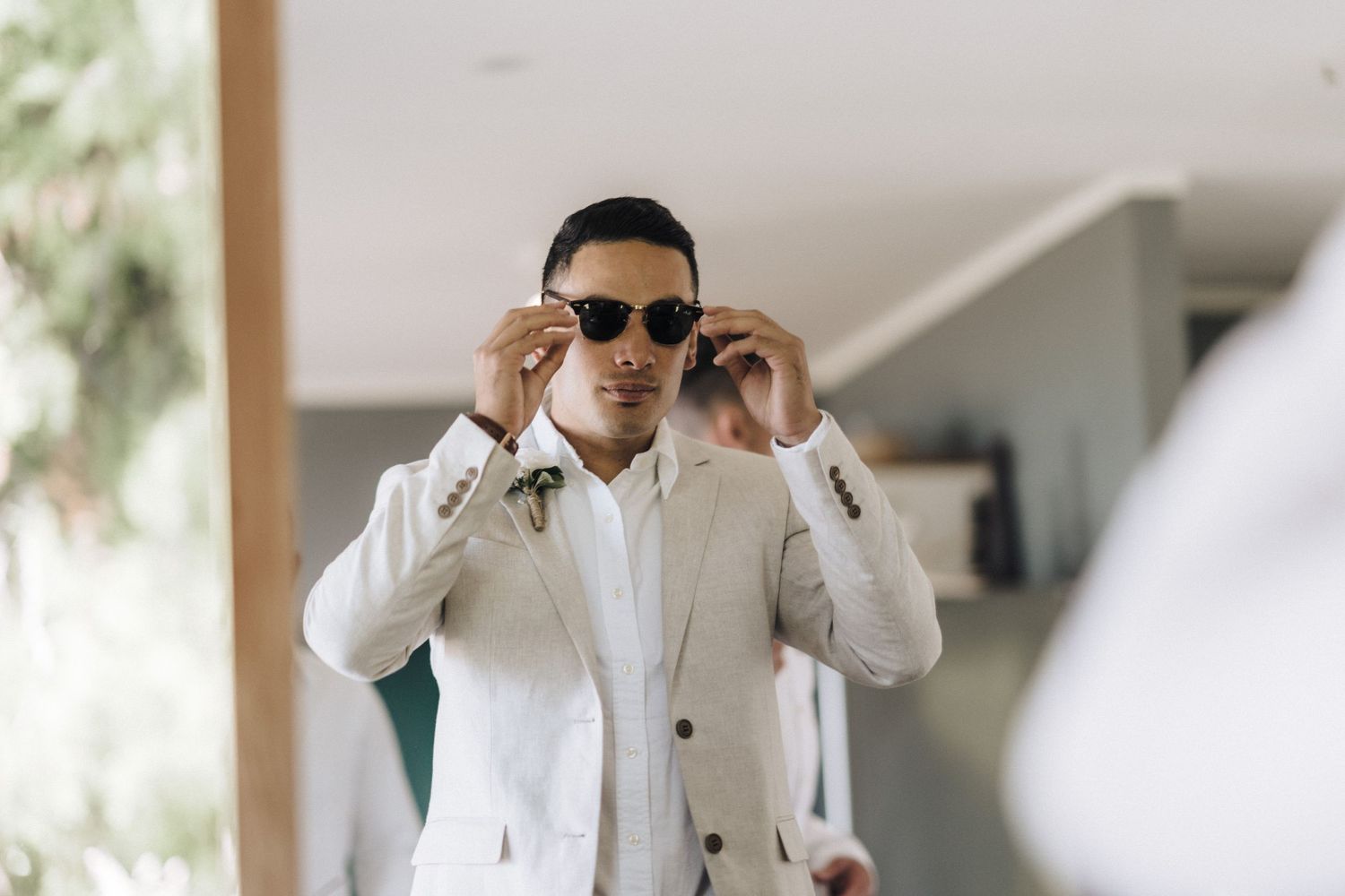 Someone wearing a white suit jacket and sunglasses adjusts their frames while standing indoors.
