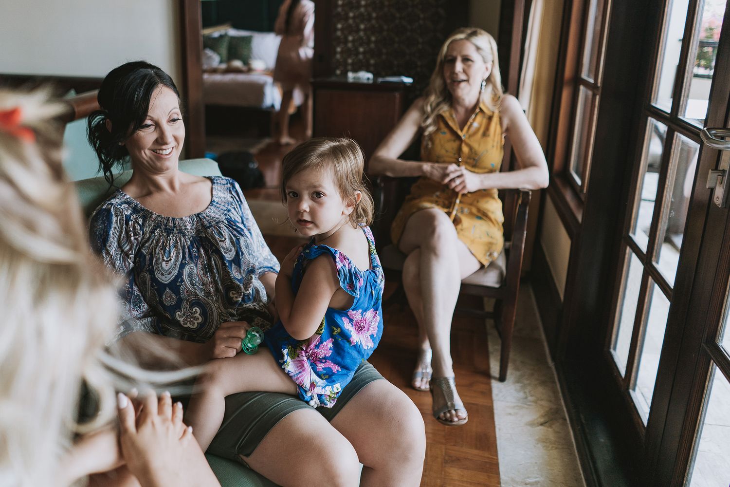Group of women relaxing and chatting by large windows during wedding preparations.