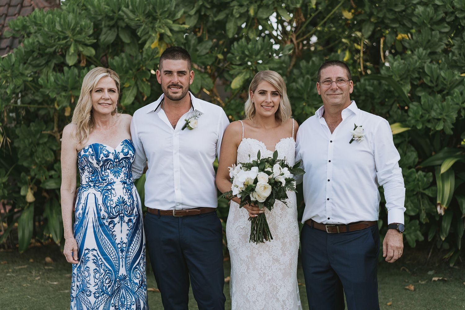 A wedding party poses together outdoors against lush greenery wearing formal attire.