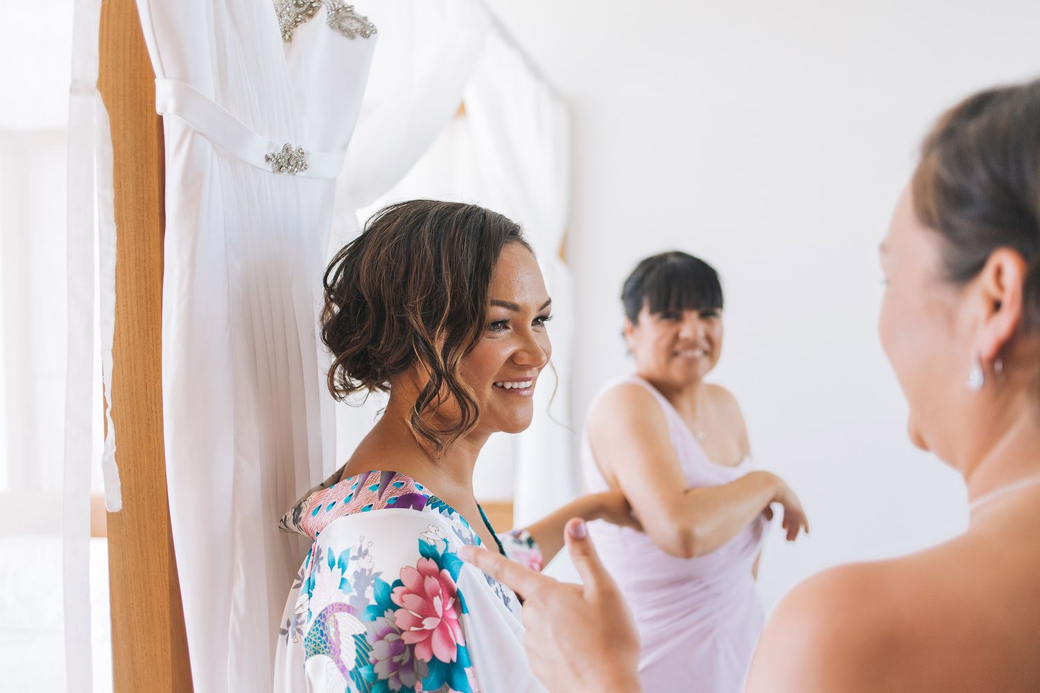 Getting ready scene shows group helping with wedding attire in a sunlit room with floral robes.