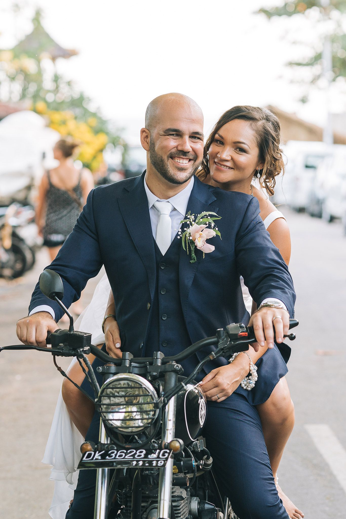 A couple in formal wedding attire poses together on a vintage motorcycle with boutonniere and bridal details.