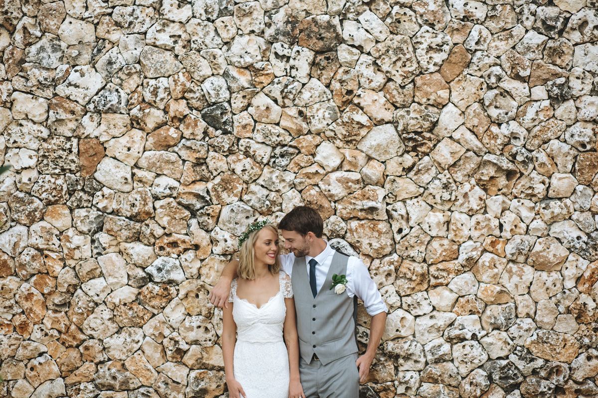 A couple in wedding attire poses against a textured stone wall backdrop.