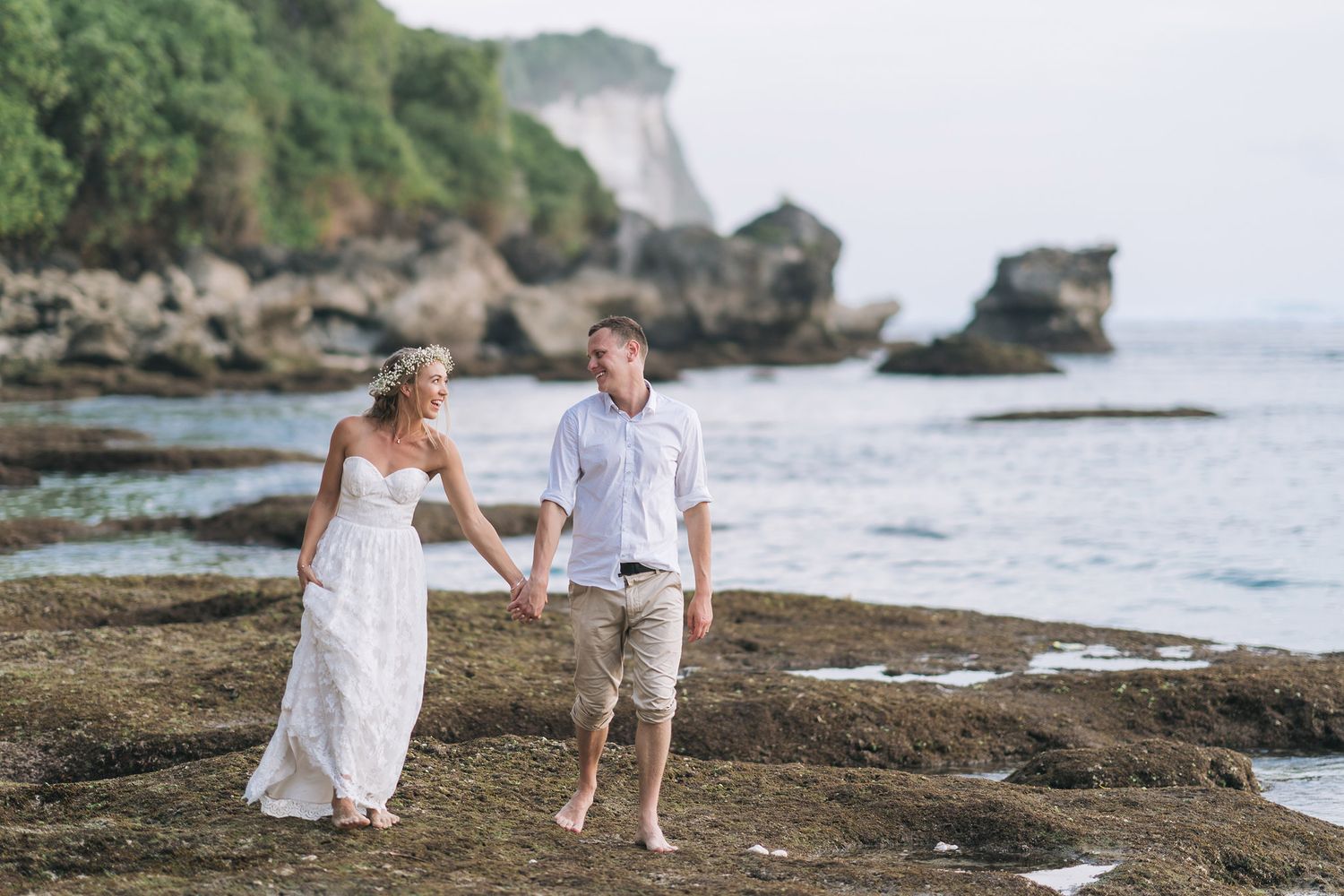 Couple walking barefoot along rocky coastline with ocean cliffs in background.