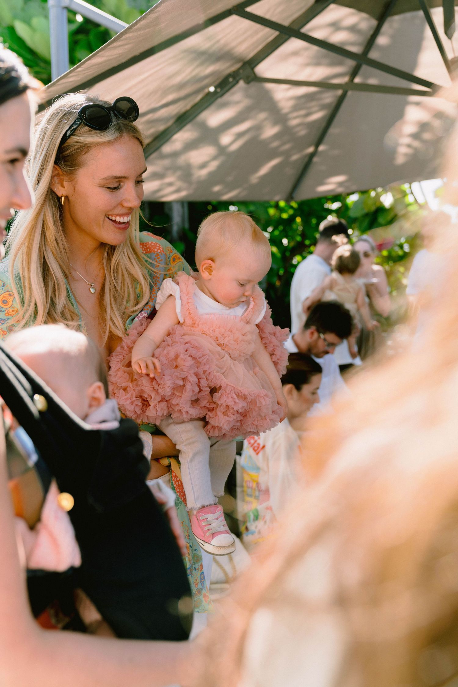 Baby in a pink tulle dress being held at an outdoor gathering under a canopy on a sunny day.