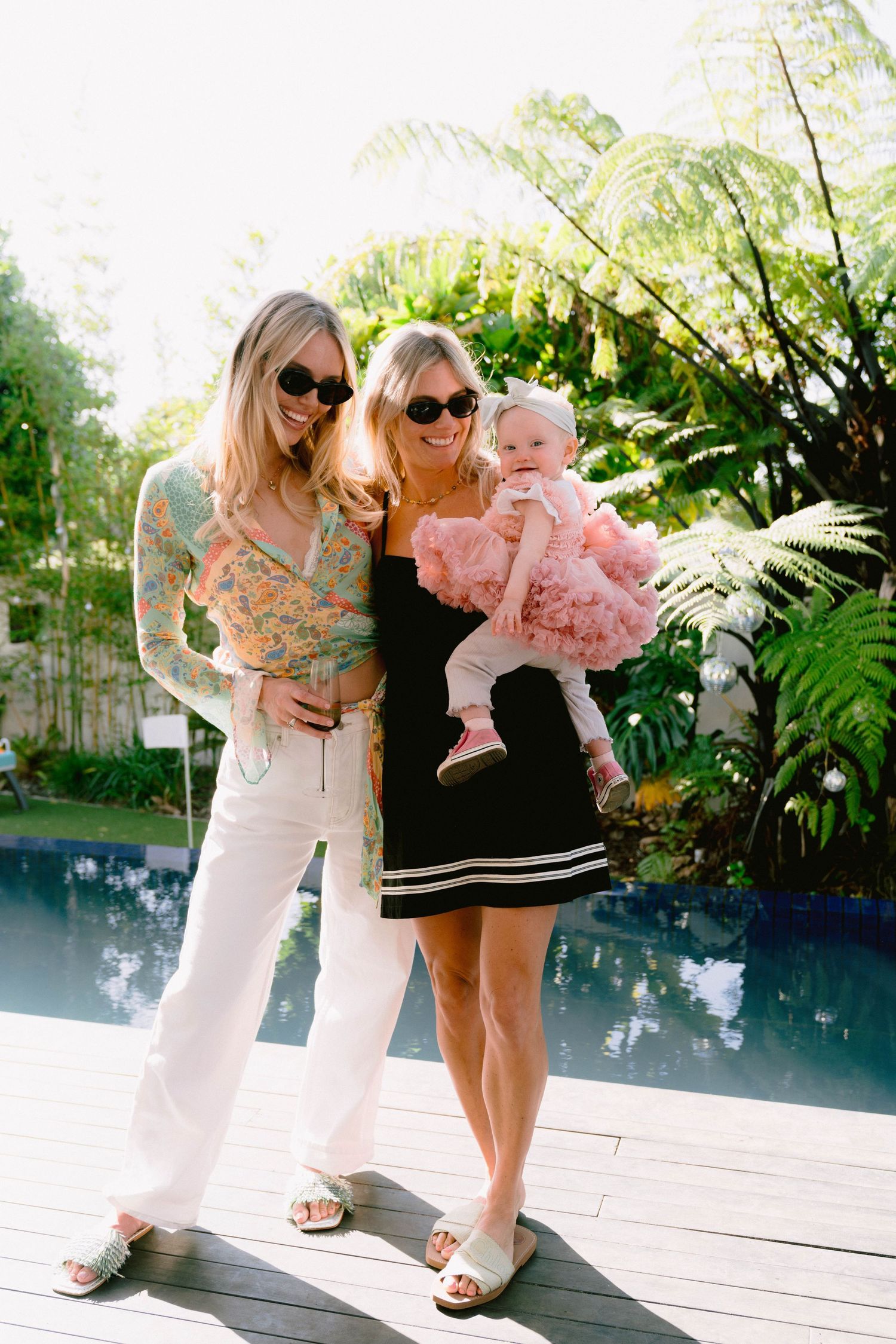 Two women wearing summer dresses and sunglasses standing near a pool with leafy tropical plants in background.