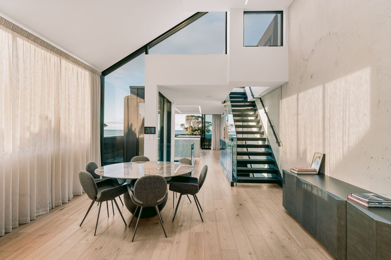 Modern open-plan dining room with round table, gray chairs, floating staircase, and floor-to-ceiling windows on light hardwood floors.