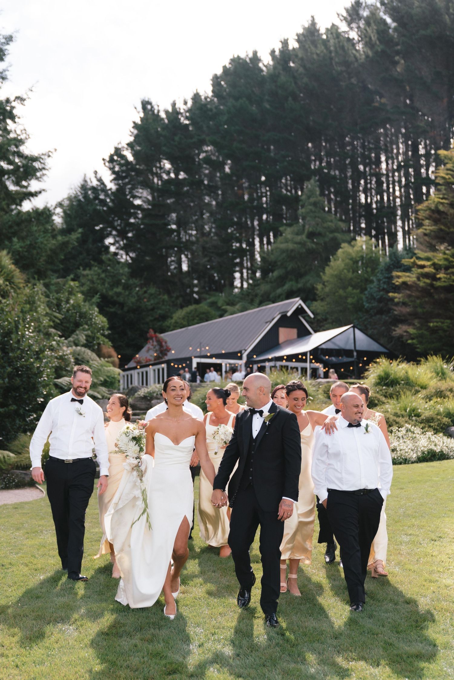 Wedding party members walking together on a lawn with evergreen trees and a rustic venue building in background.
