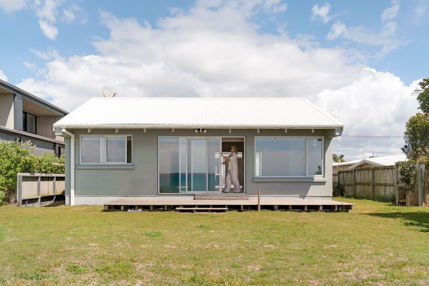 Small gray beach house with large windows and deck overlooking grassy yard on sunny day with white clouds.