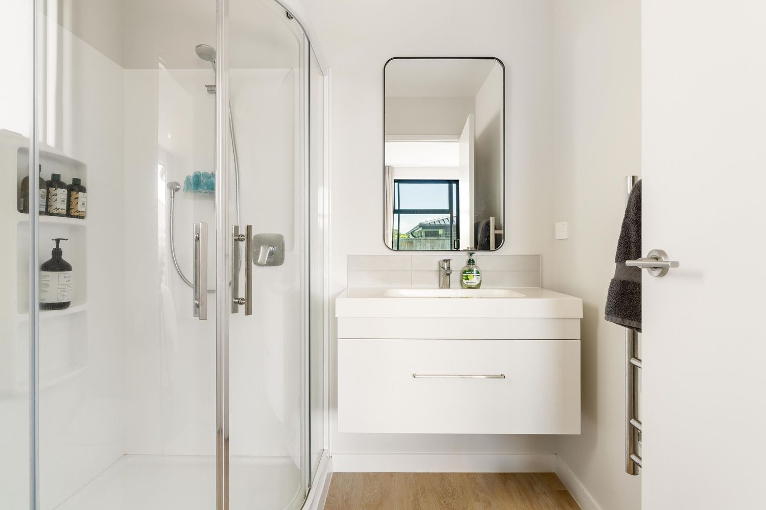 Clean white bathroom interior with floating vanity, mirror, glass shower enclosure and wooden floor.