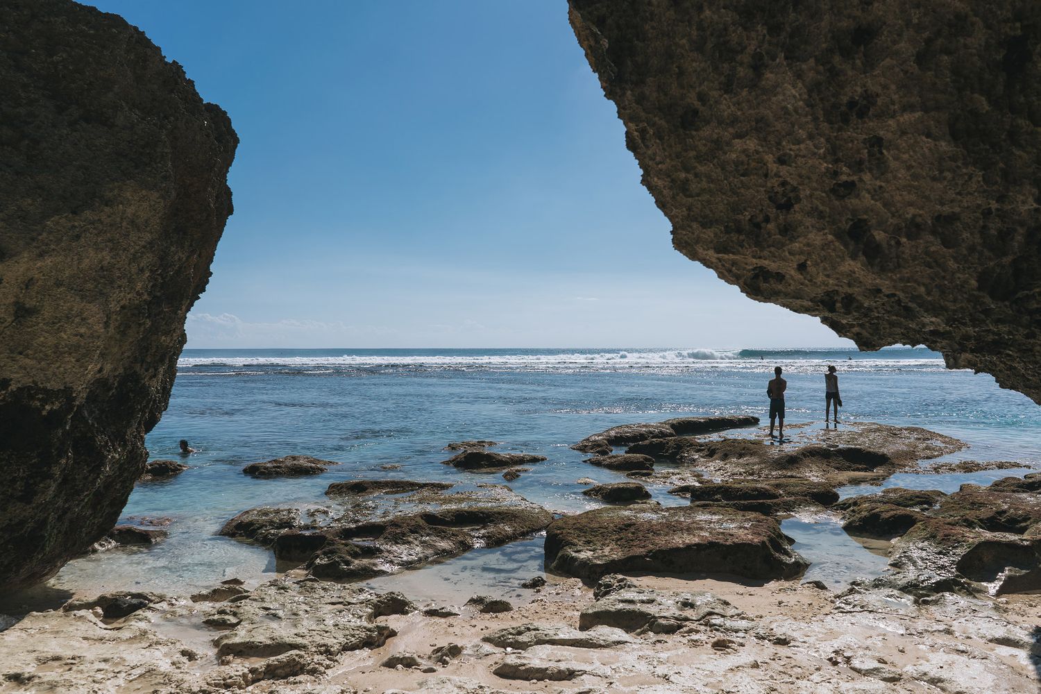 Two silhouettes stand on rocky coastline under natural stone arch formation overlooking blue ocean and distant waves.