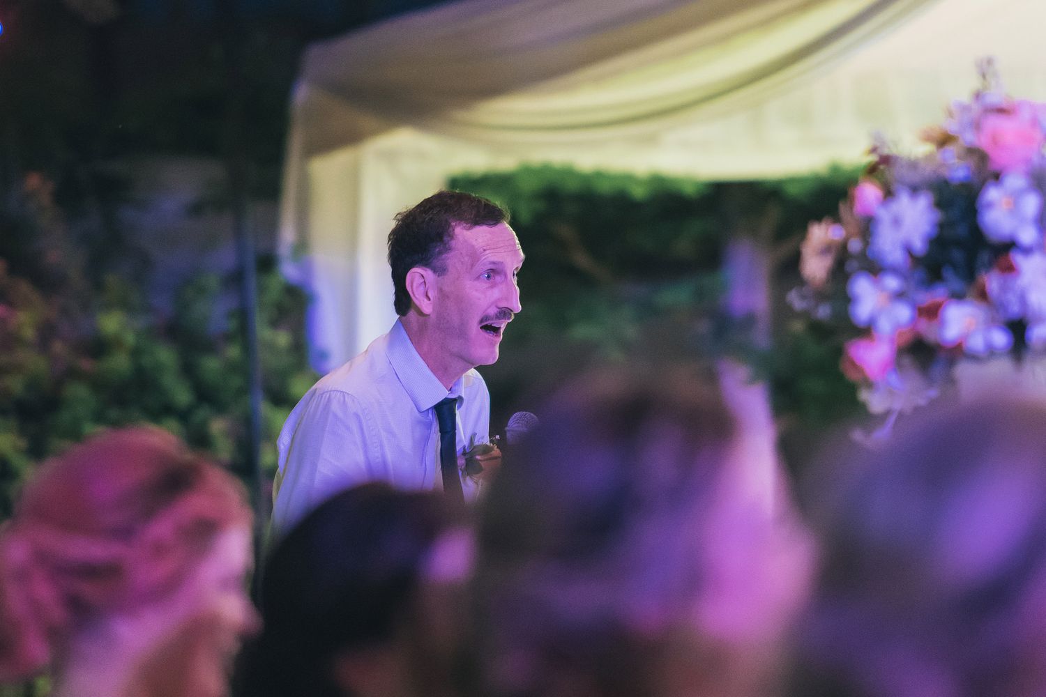 Speaker addressing wedding guests during evening reception with purple lighting and floral decor.