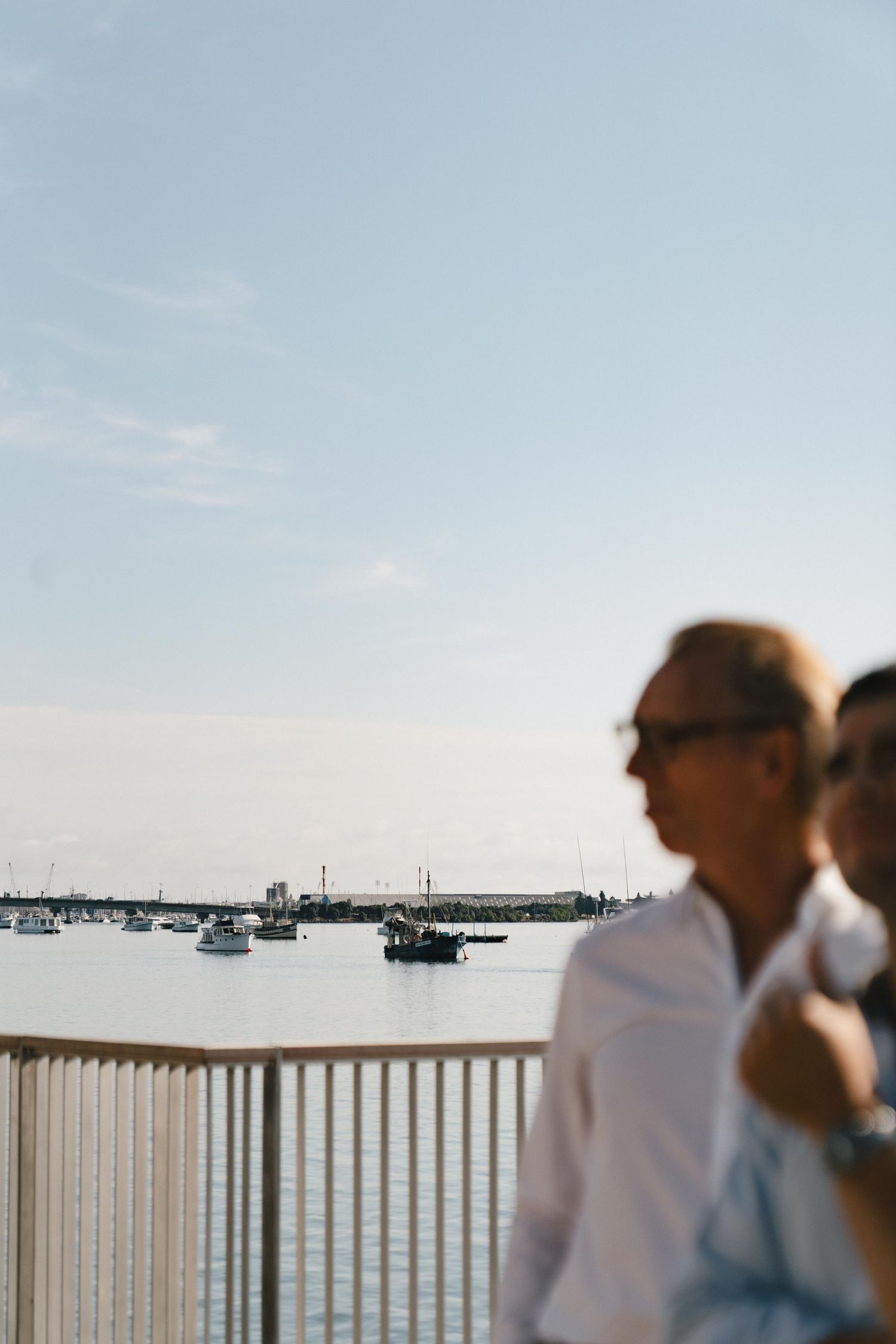 People enjoying the scenic view of boats on the water from a white railing deck on a calm day.