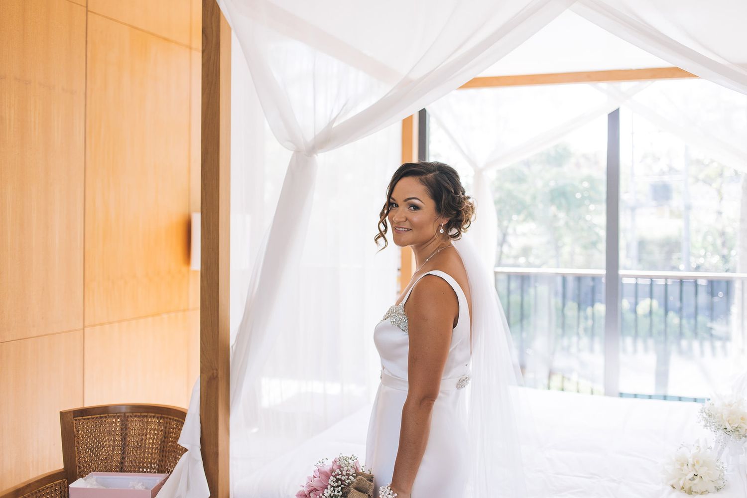 A bride in a white wedding dress poses elegantly near a window in a sunlit room with sheer curtains.