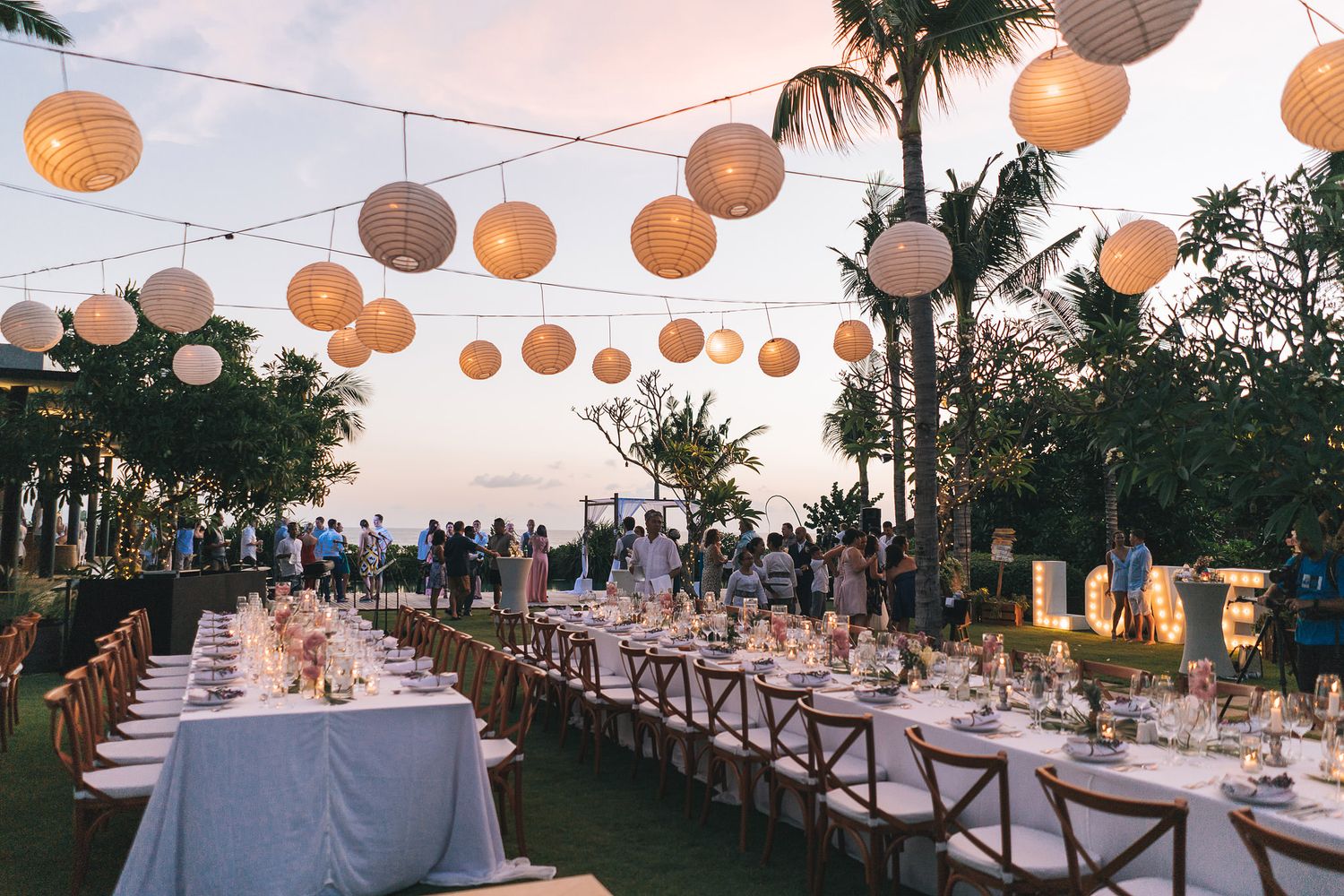 Elegant outdoor wedding reception setup with long tables under string lights and paper lanterns at sunset.