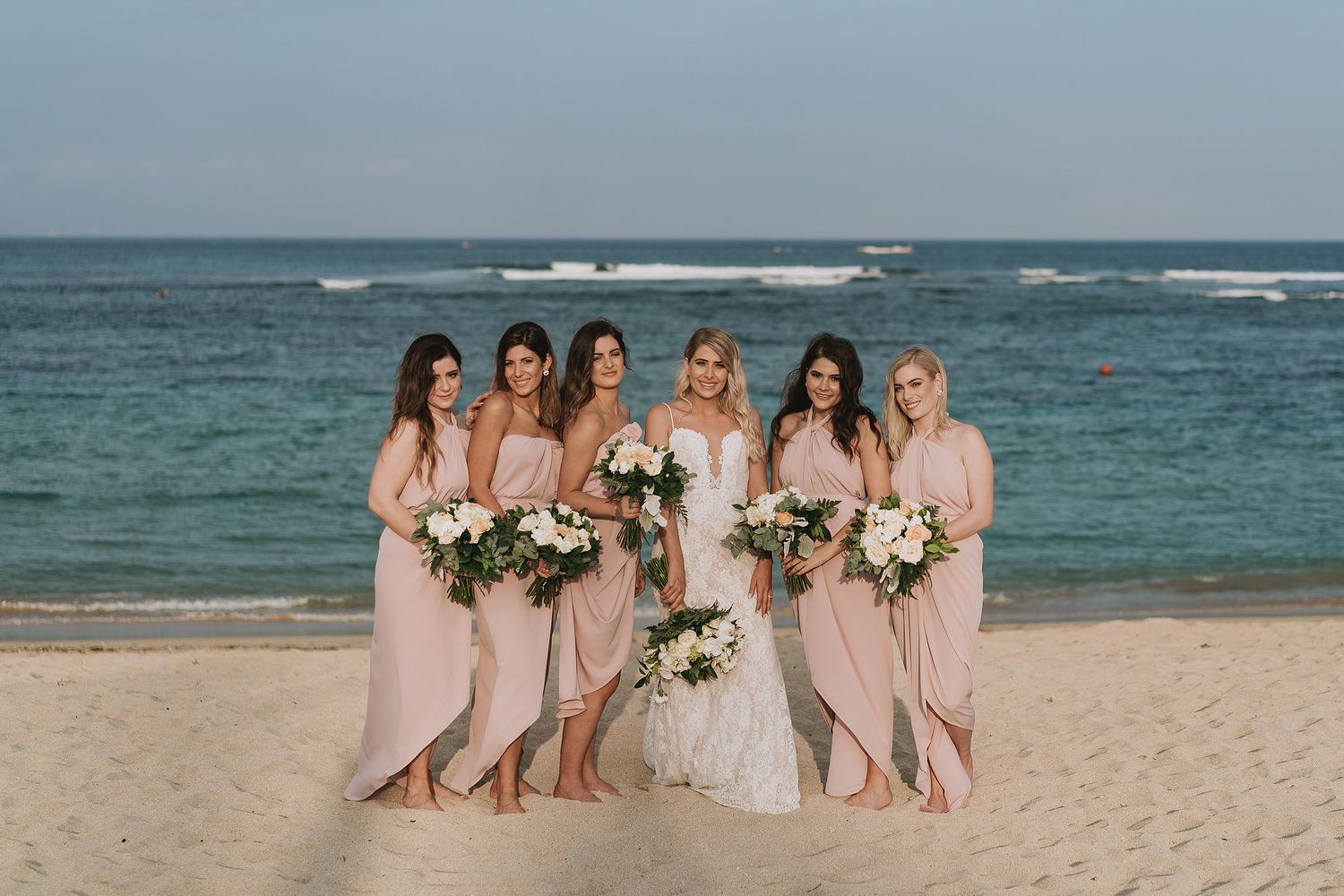 Wedding party in light pink dresses poses for photos on a scenic beach with ocean waves in the background.