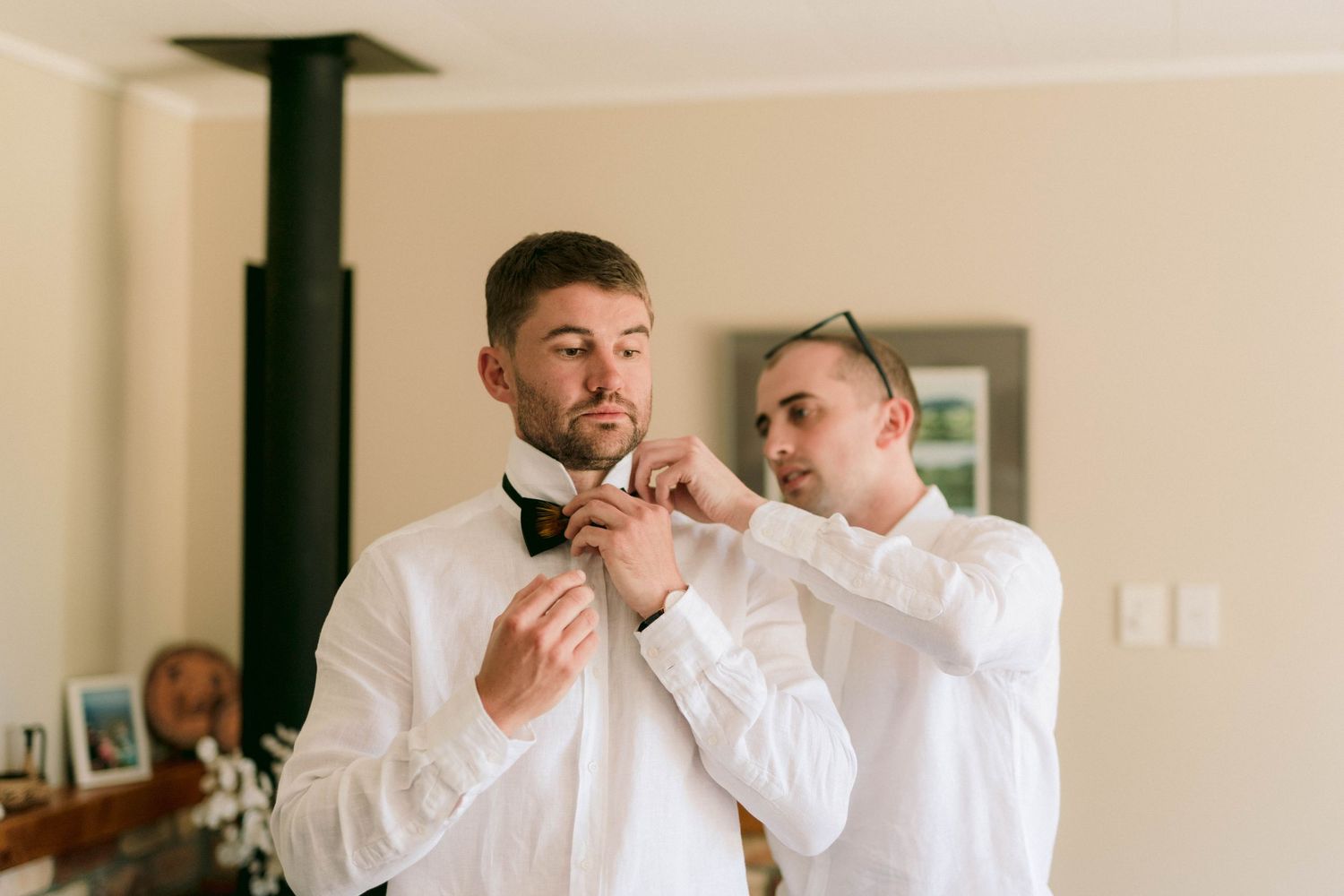 Two people in white dress shirts help fix a bowtie while getting ready in a bright interior room.
