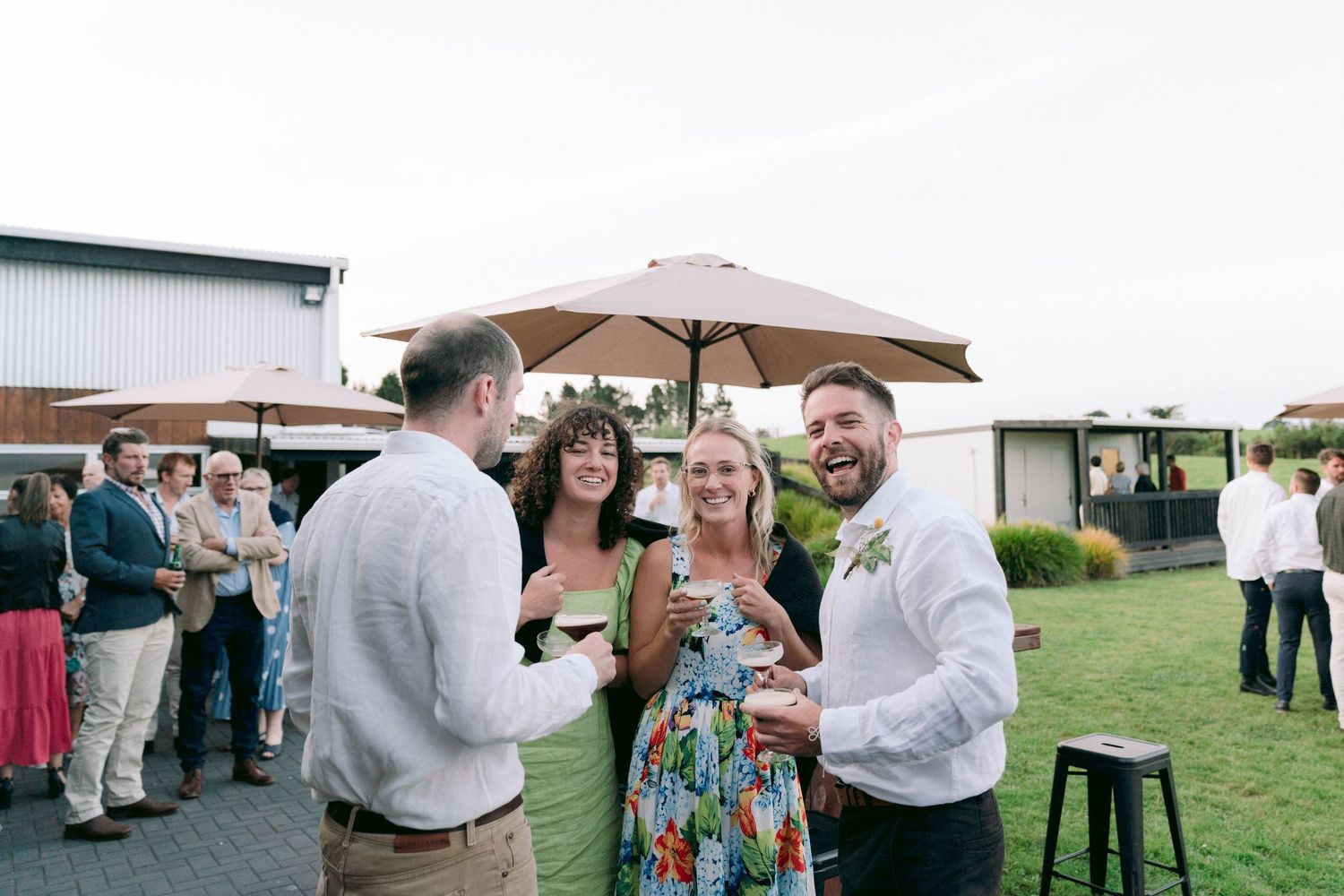 Wedding guests socializing outdoors under a beige umbrella during cocktail hour.