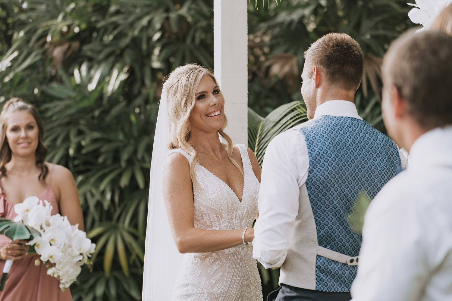 A wedding ceremony takes place outdoors with tropical foliage in the background.