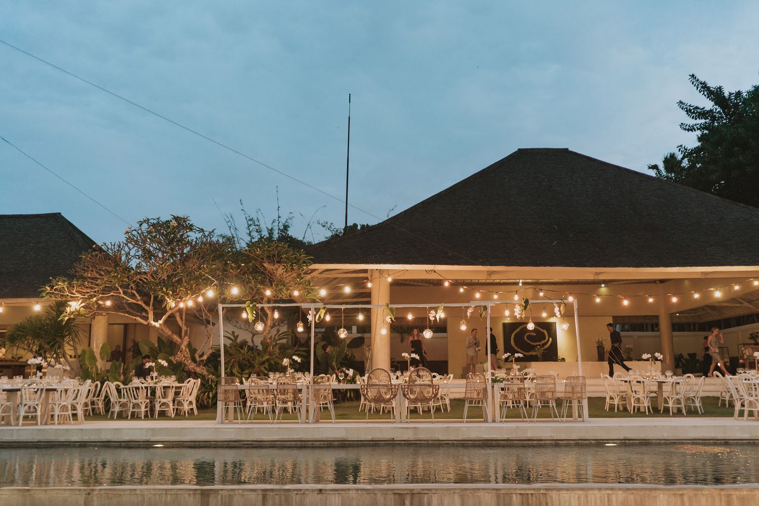 Outdoor reception venue with string lights and white tables set up under evening sky.
