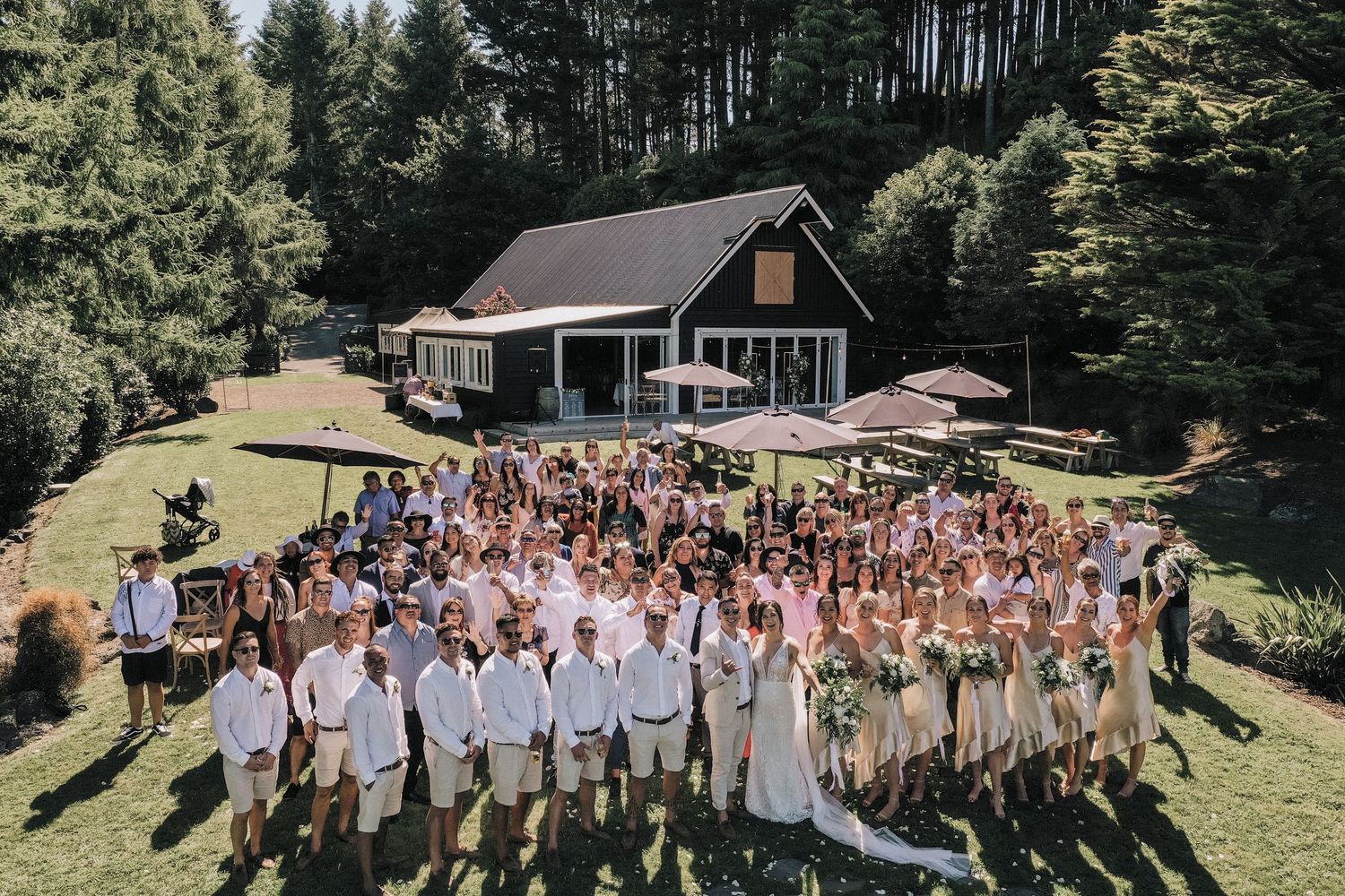 Aerial view of large wedding group photo in front of modern cabin surrounded by pine forest.