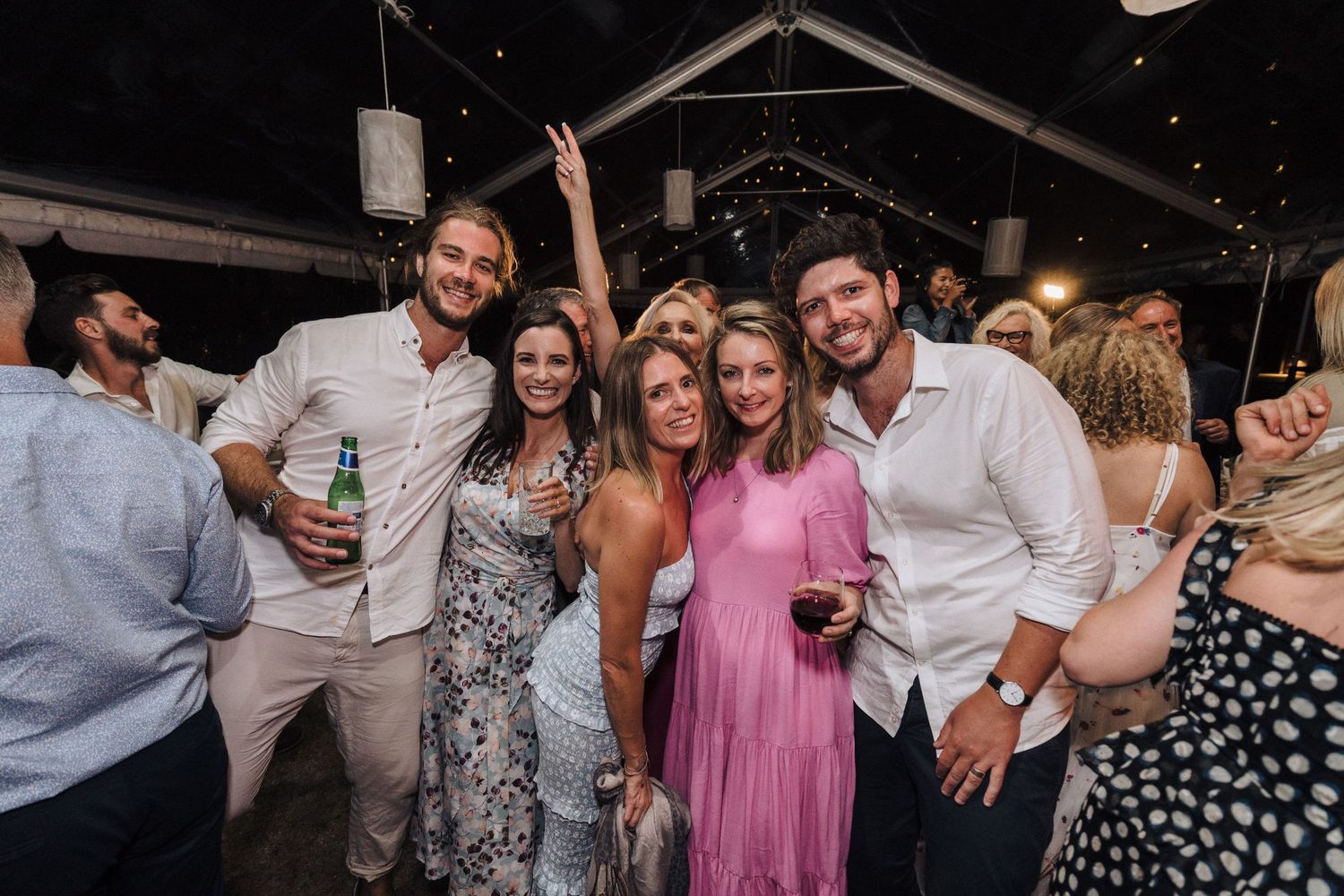 A group of wedding guests celebrate at an outdoor reception under string lights.