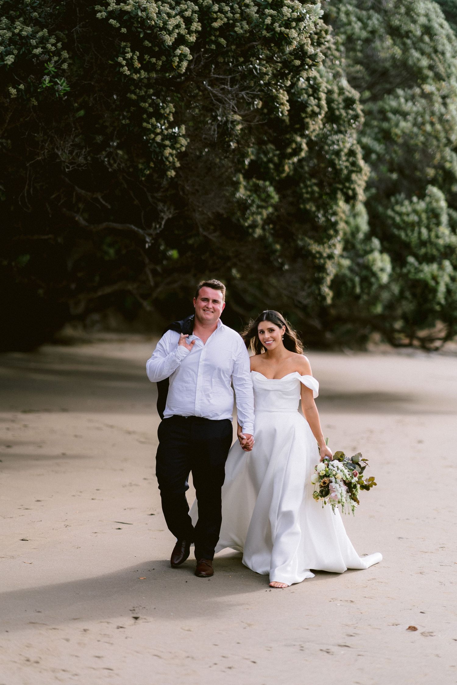 Newlyweds walking hand in hand on sandy beach path surrounded by lush greenery.