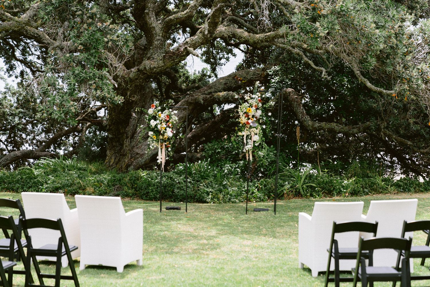 An outdoor wedding ceremony setup with white chairs and floral arch arrangements under a majestic tree.
