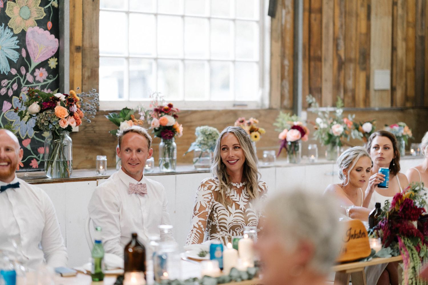 Wedding guests smile and celebrate during a rustic barn reception with floral centerpieces and natural light.