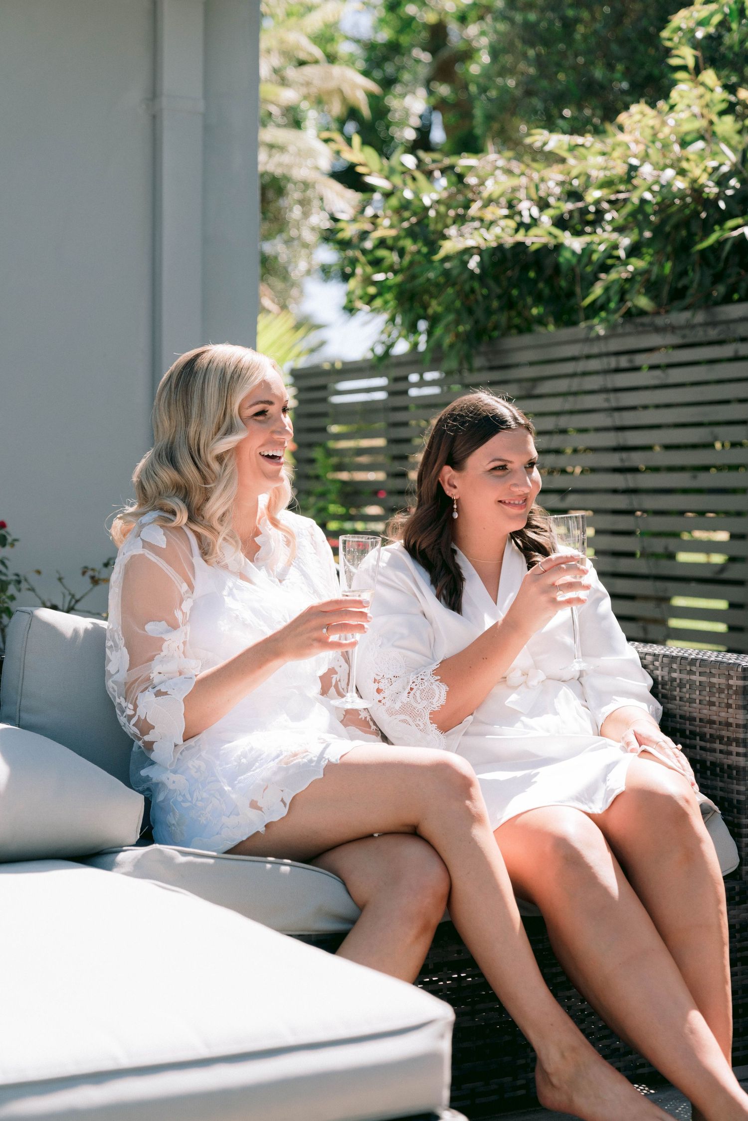 Two bridesmaids in white robes sharing laughter while lounging on outdoor furniture.