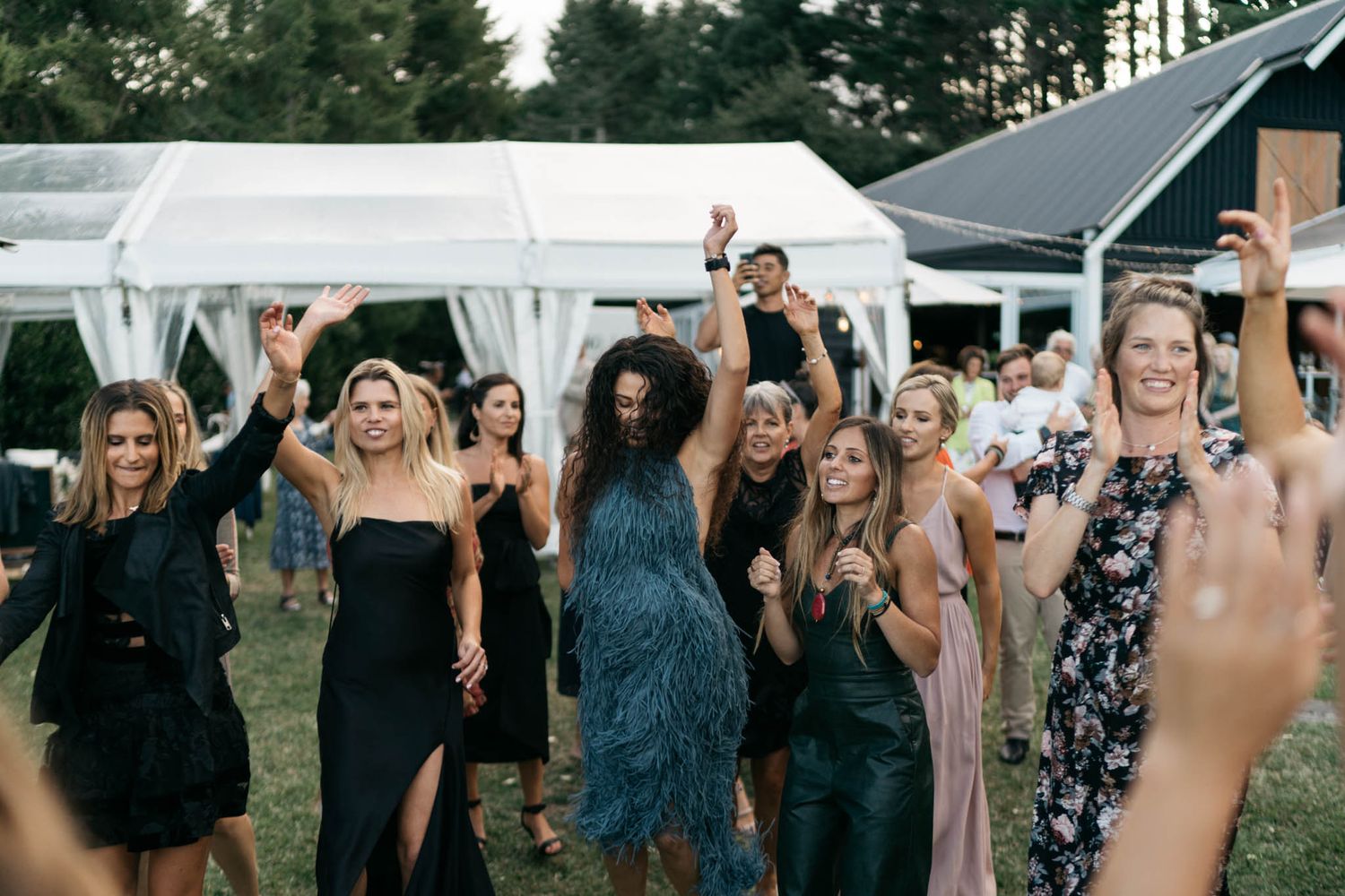 Wedding guests dance joyfully under a white tent during an outdoor reception celebration.