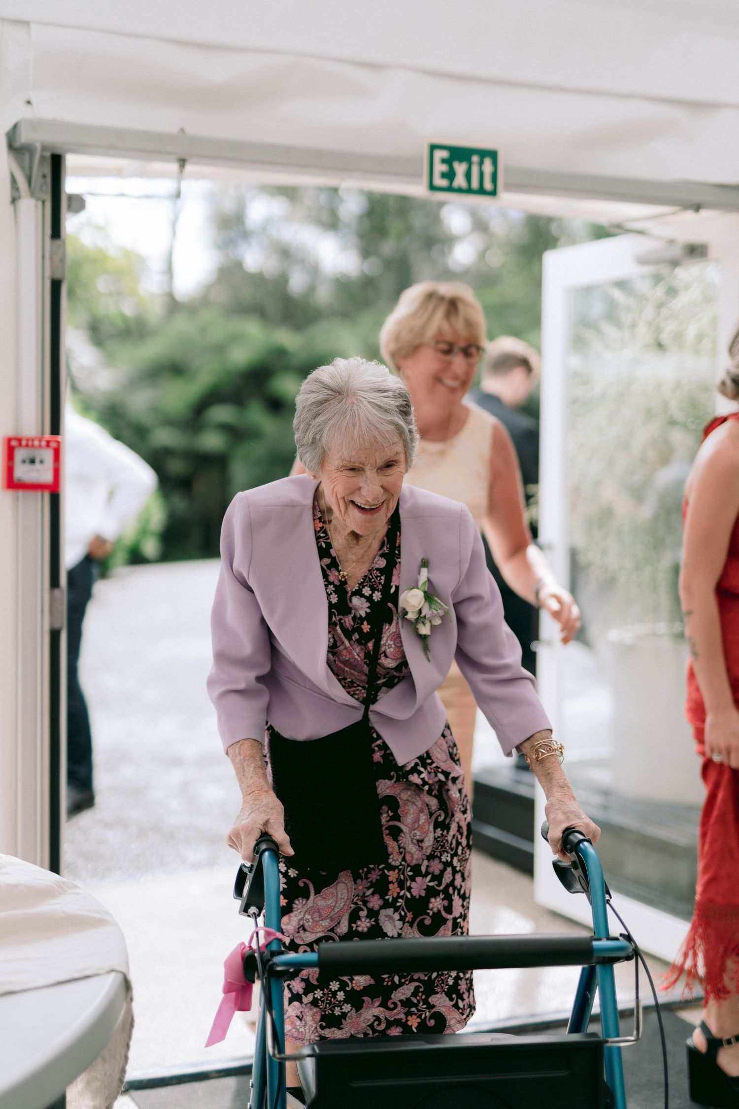 An elderly guest using a walker arrives at an outdoor event venue with an exit sign visible.