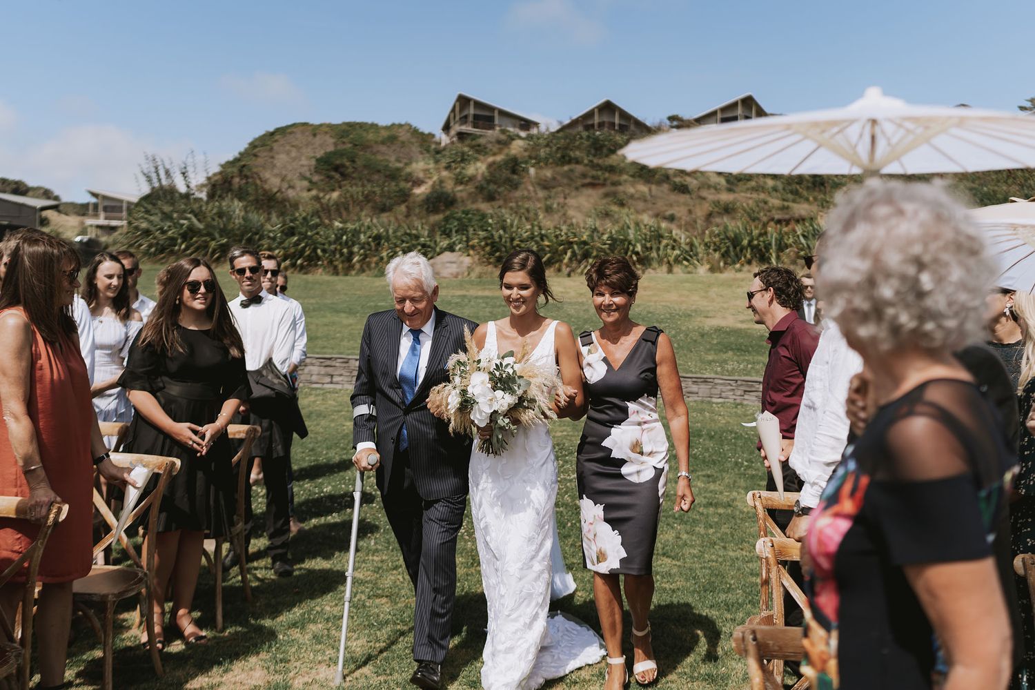 Wedding guests gather outdoors on a grassy hillside under blue skies during a ceremony on a sunny day.