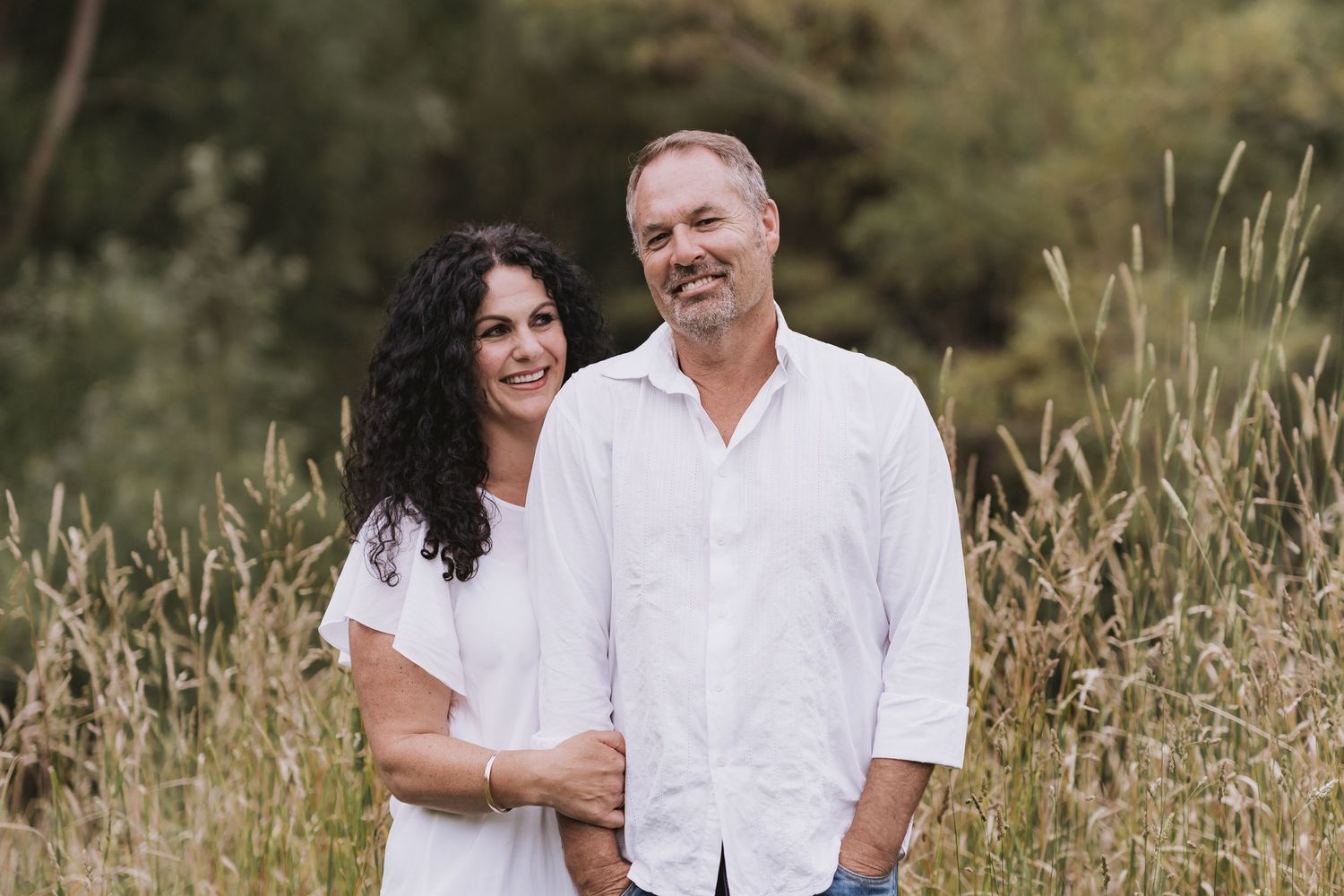 Two people wearing white shirts embrace in a scenic grassy meadow at golden hour.