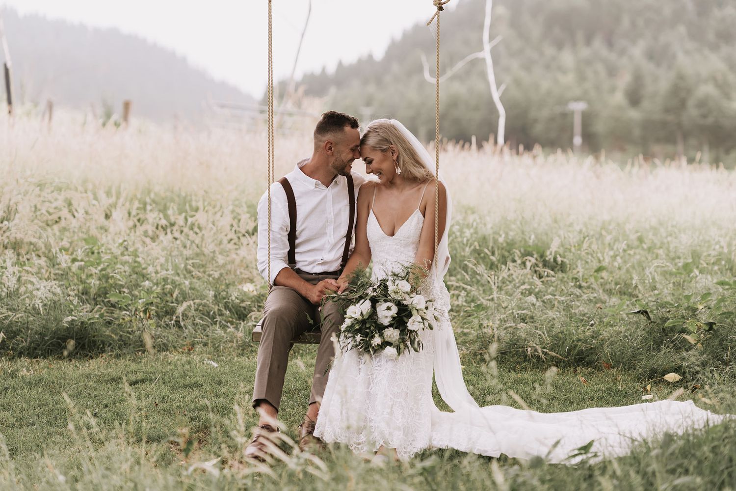 Couple shares intimate moment on grassy hillside in elegant wedding attire with mountain backdrop and bouquet.