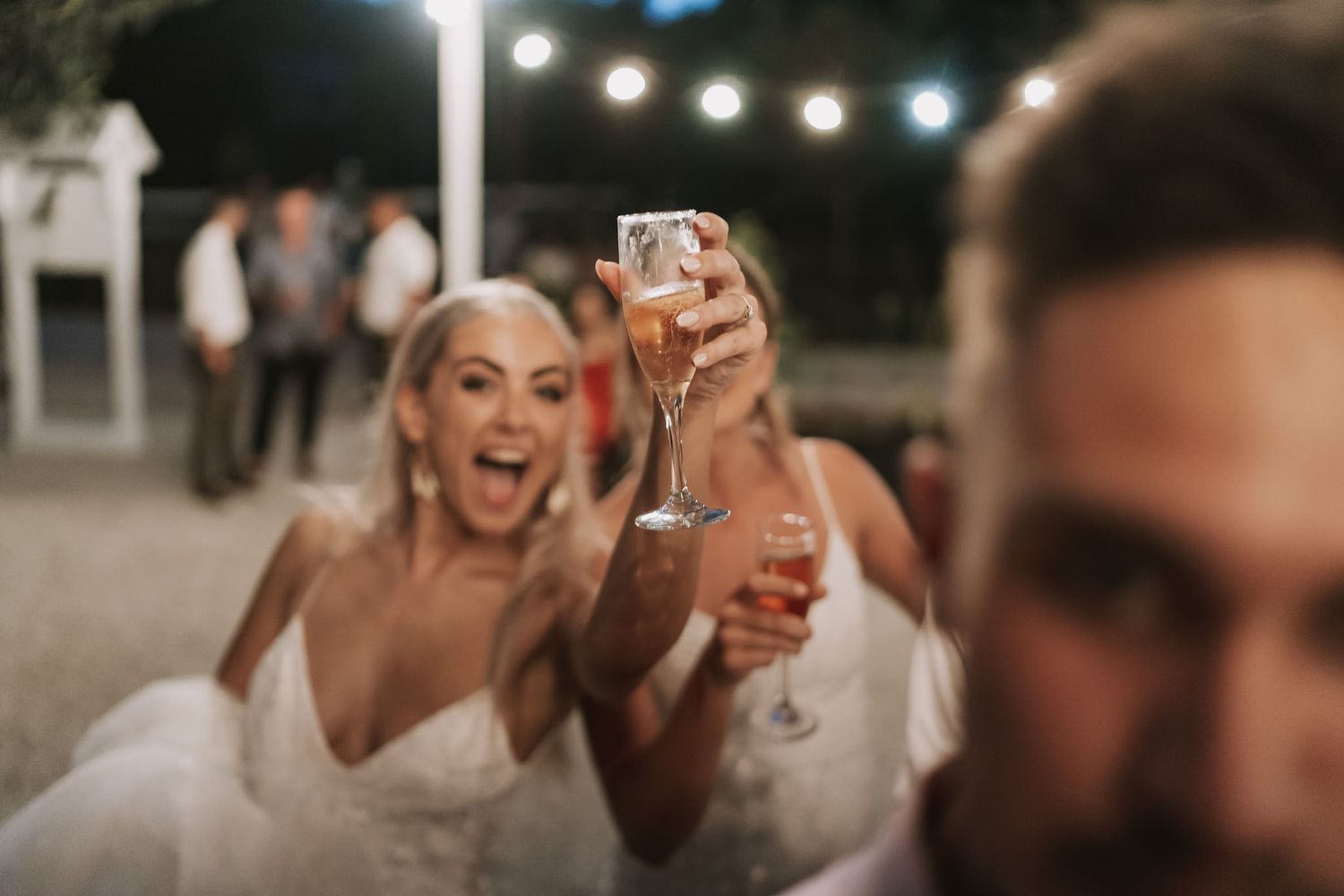 A lively wedding reception celebration with people toasting drinks under string lights at night.