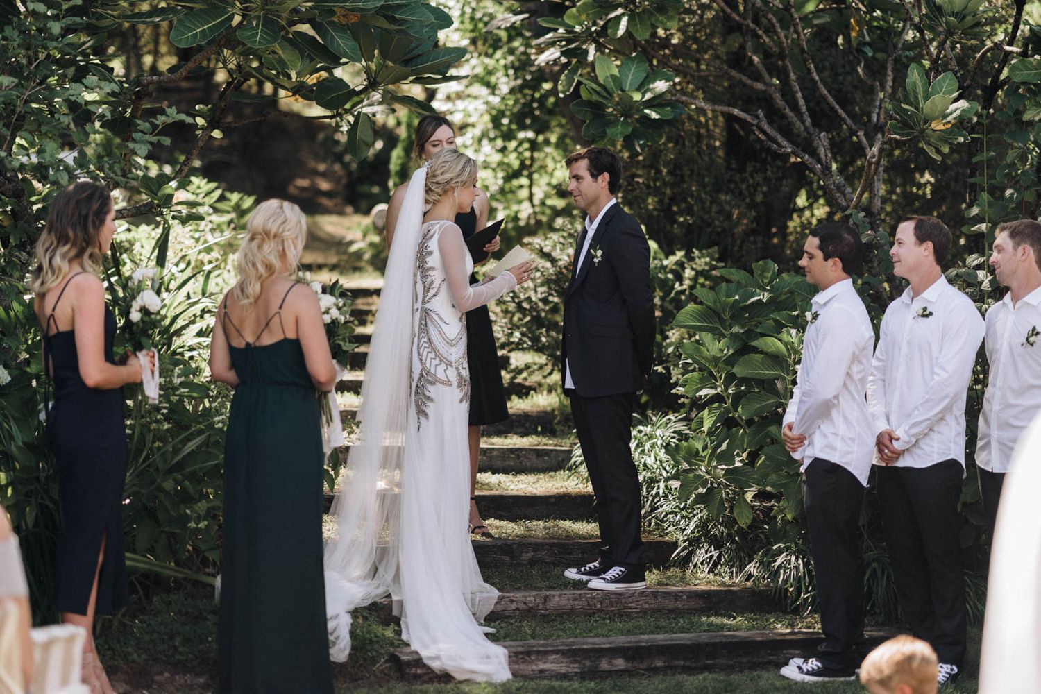 An outdoor wedding ceremony takes place in a garden setting with lush greenery and trees in the background.