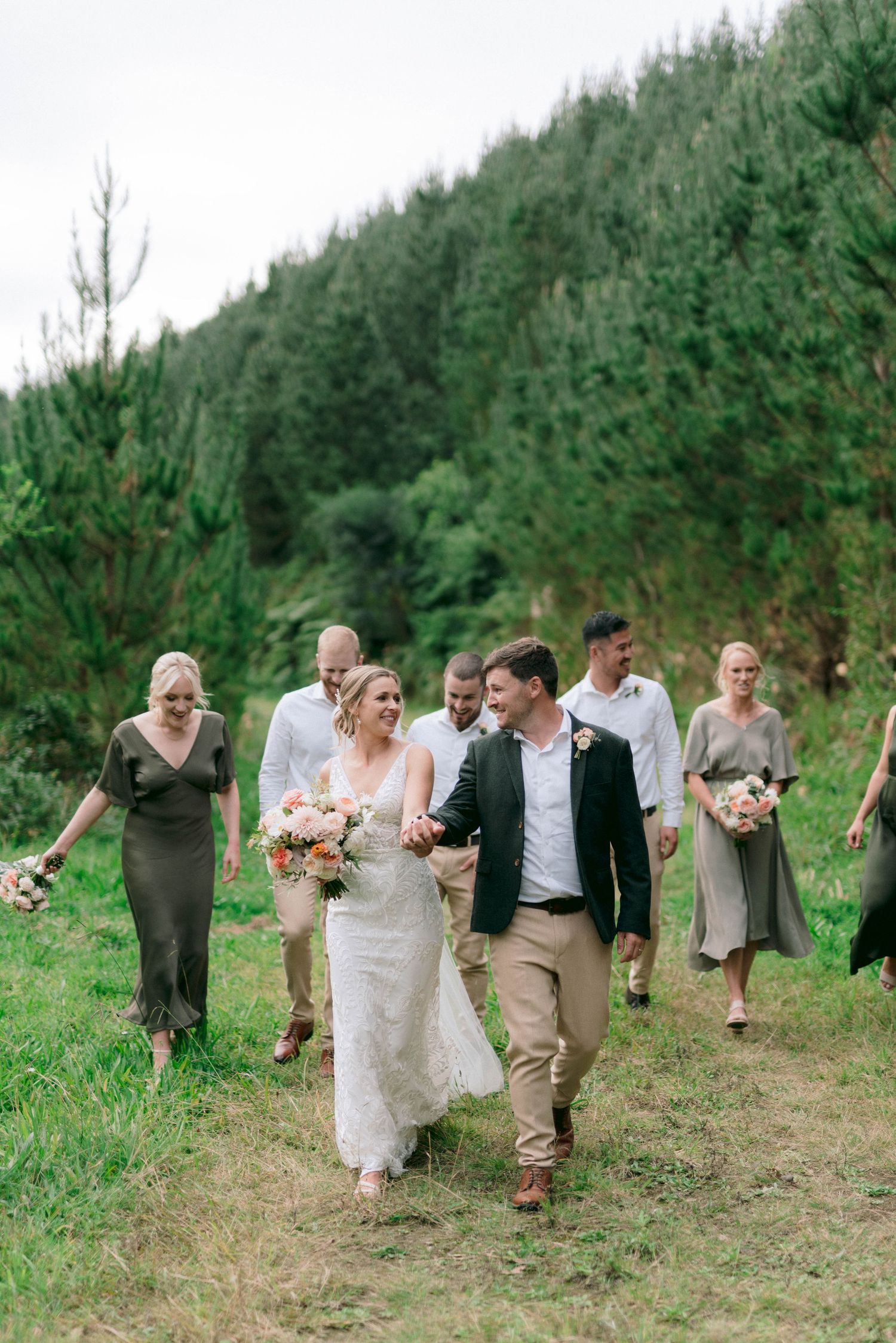 A wedding party walks along a forest path with evergreen trees in the background on a cloudy day.